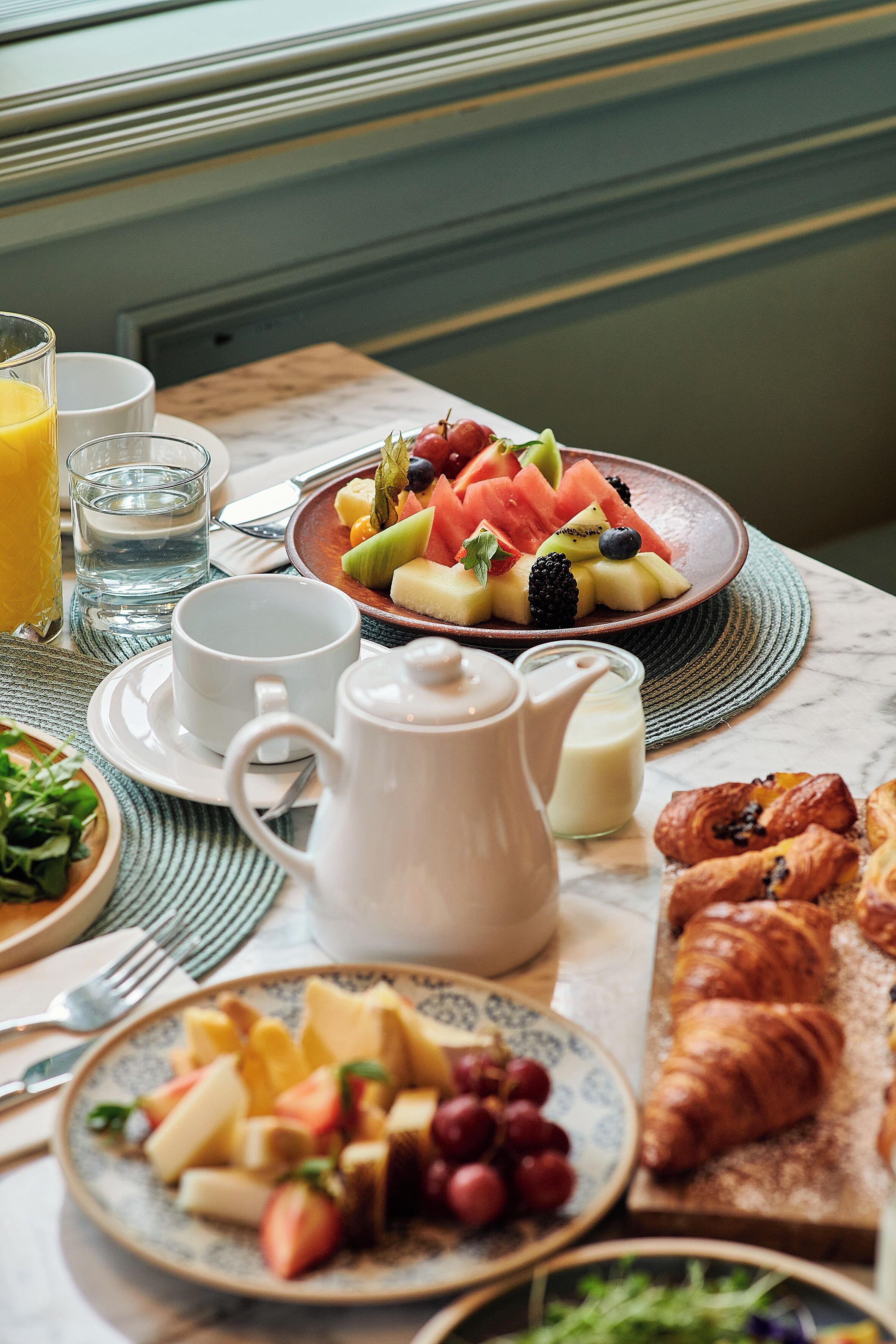 A breakfast setting with plates of fruit, croissants, and a teapot on a marble table.