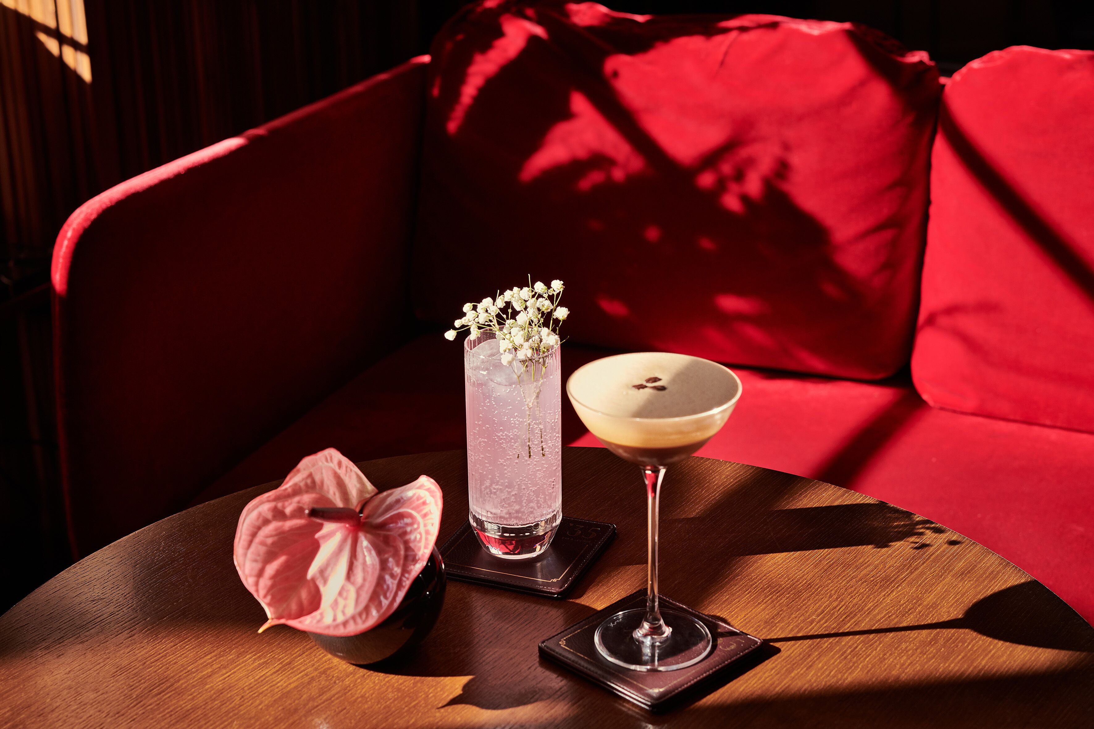 A table with a drink, a glass vase with flowers, and a pink flower on a wooden table in a room with a red couch.