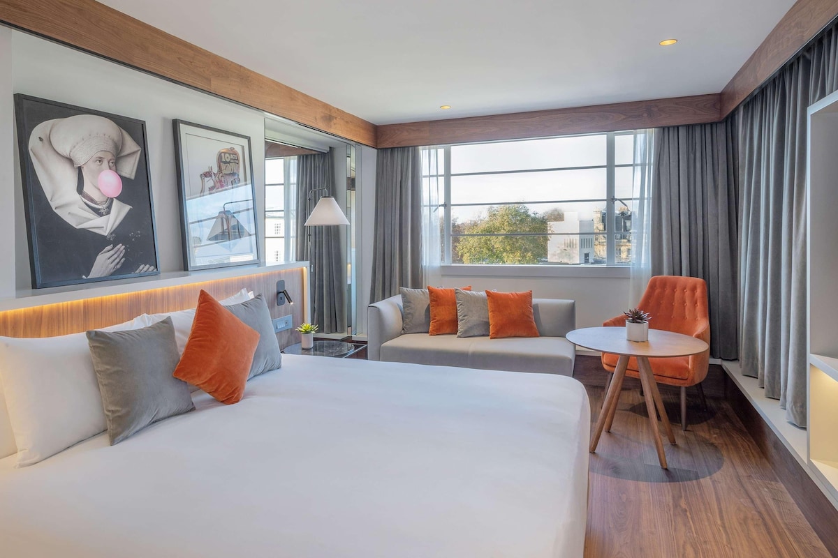 A hotel room with a large bed, two smaller sofas, a round table, and a view of the city through large windows.