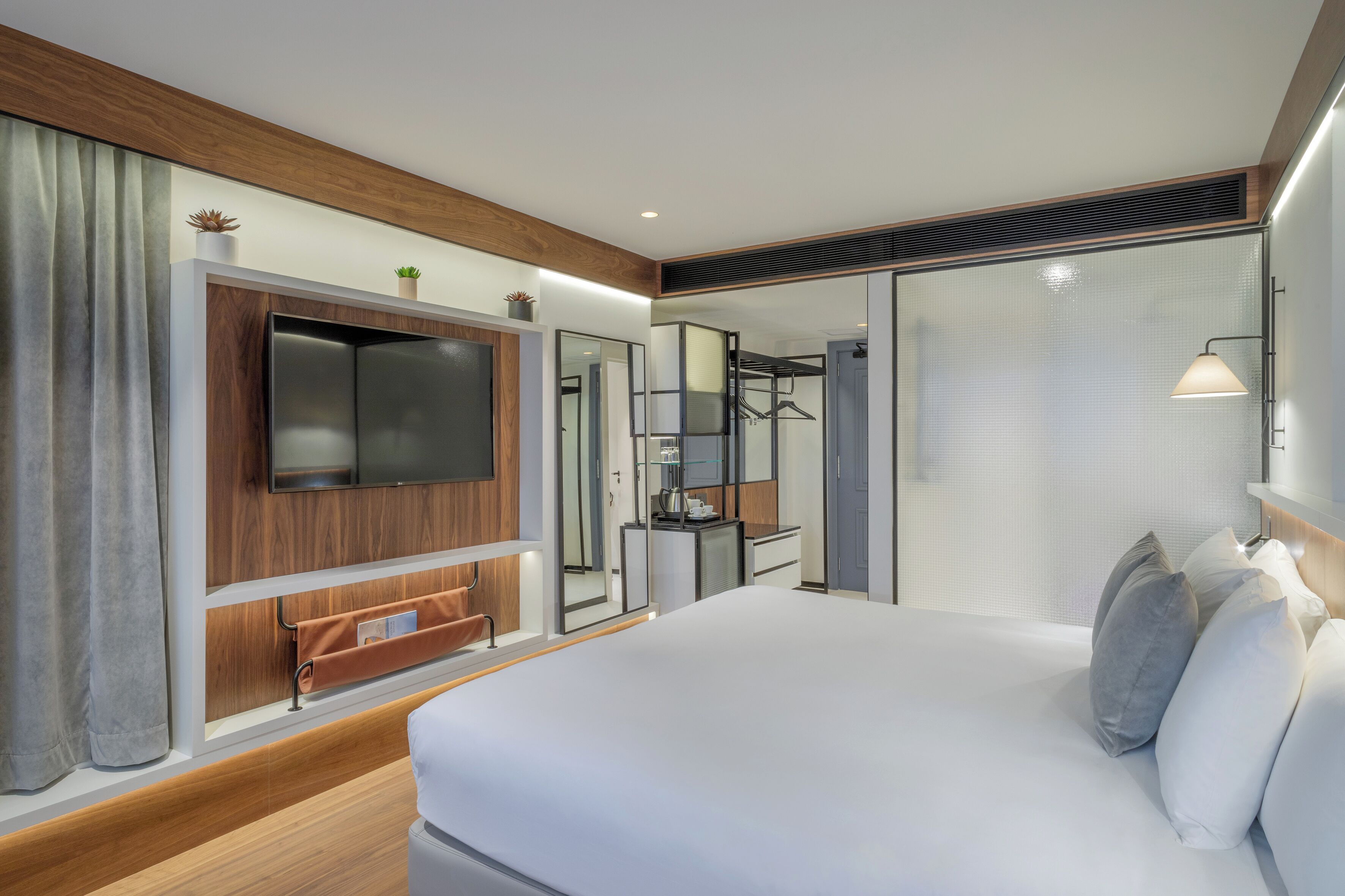 Una habitación de hotel moderna con una cama grande, un televisor de pantalla plana montado en un panel de madera, un escritorio pequeño con una silla y un espejo.
