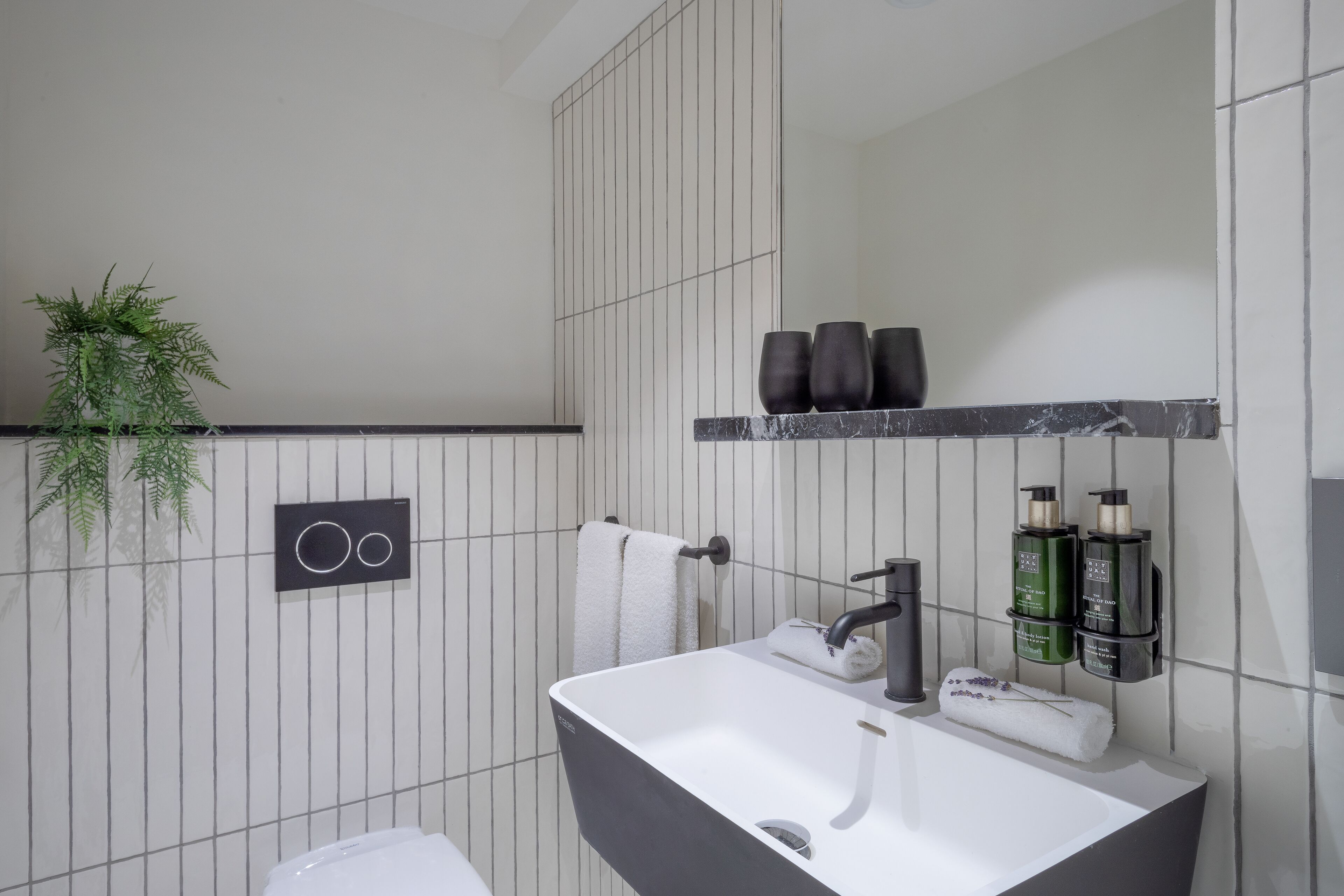 Un baño moderno con un lavamanos blanco, un grifo negro y un toallero con dos toallas. Hay dos botellas verdes en una repisa sobre el lavamanos.