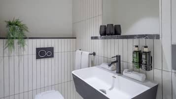 Un baño moderno con un lavamanos blanco, un grifo negro y un toallero con dos toallas. Hay dos botellas verdes en una repisa sobre el lavamanos.