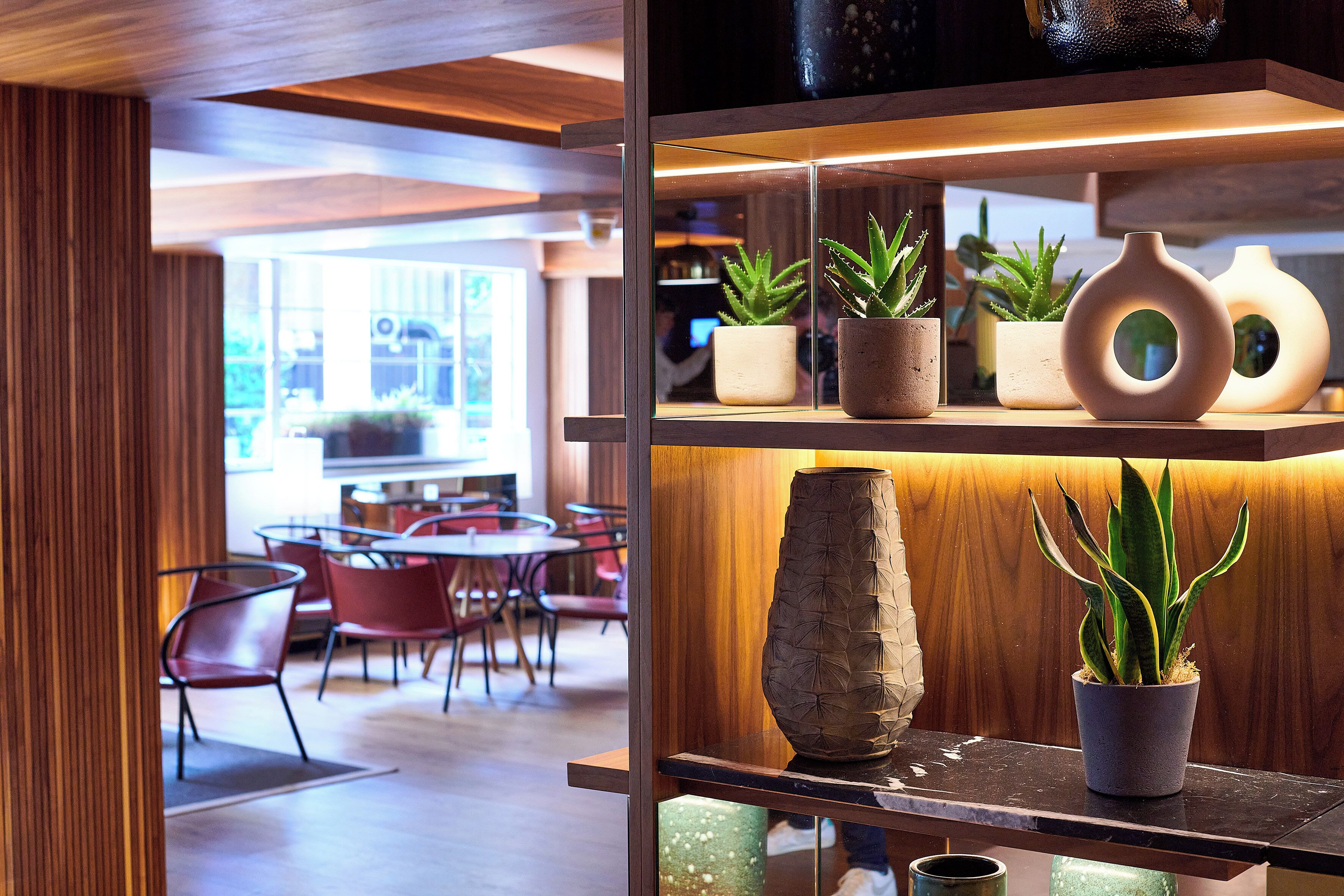 A modern interior with a wooden reception desk, a seating area with red chairs, and a display of potted plants and vases.