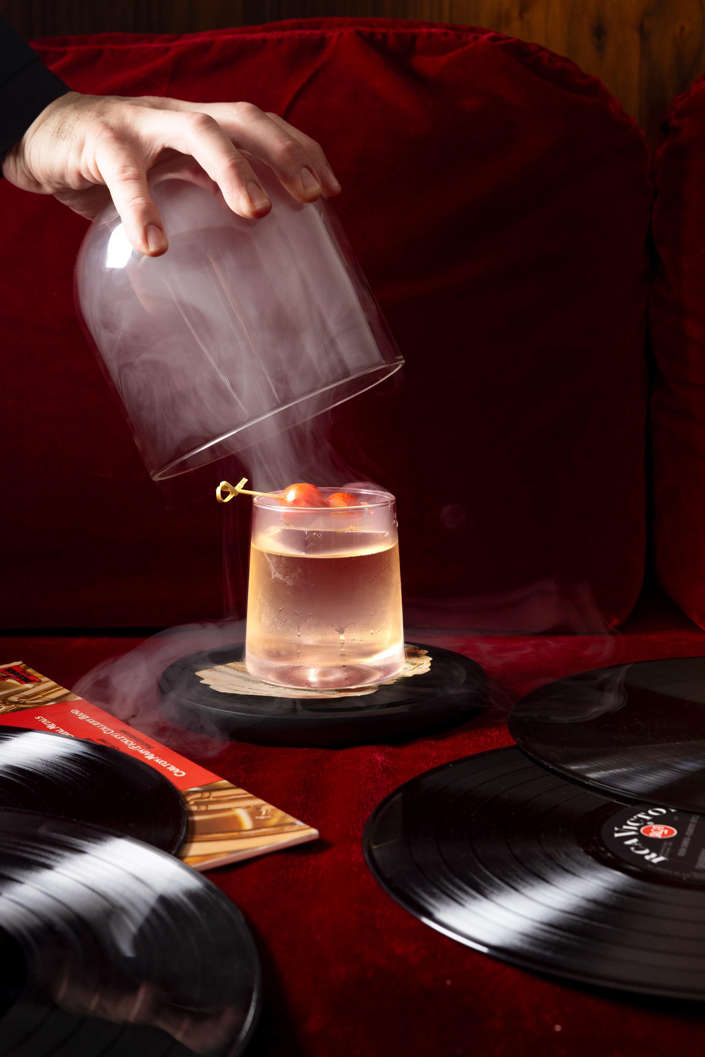 A person pouring a drink into a glass on a coaster. There are vinyl records and a red cloth in the background.