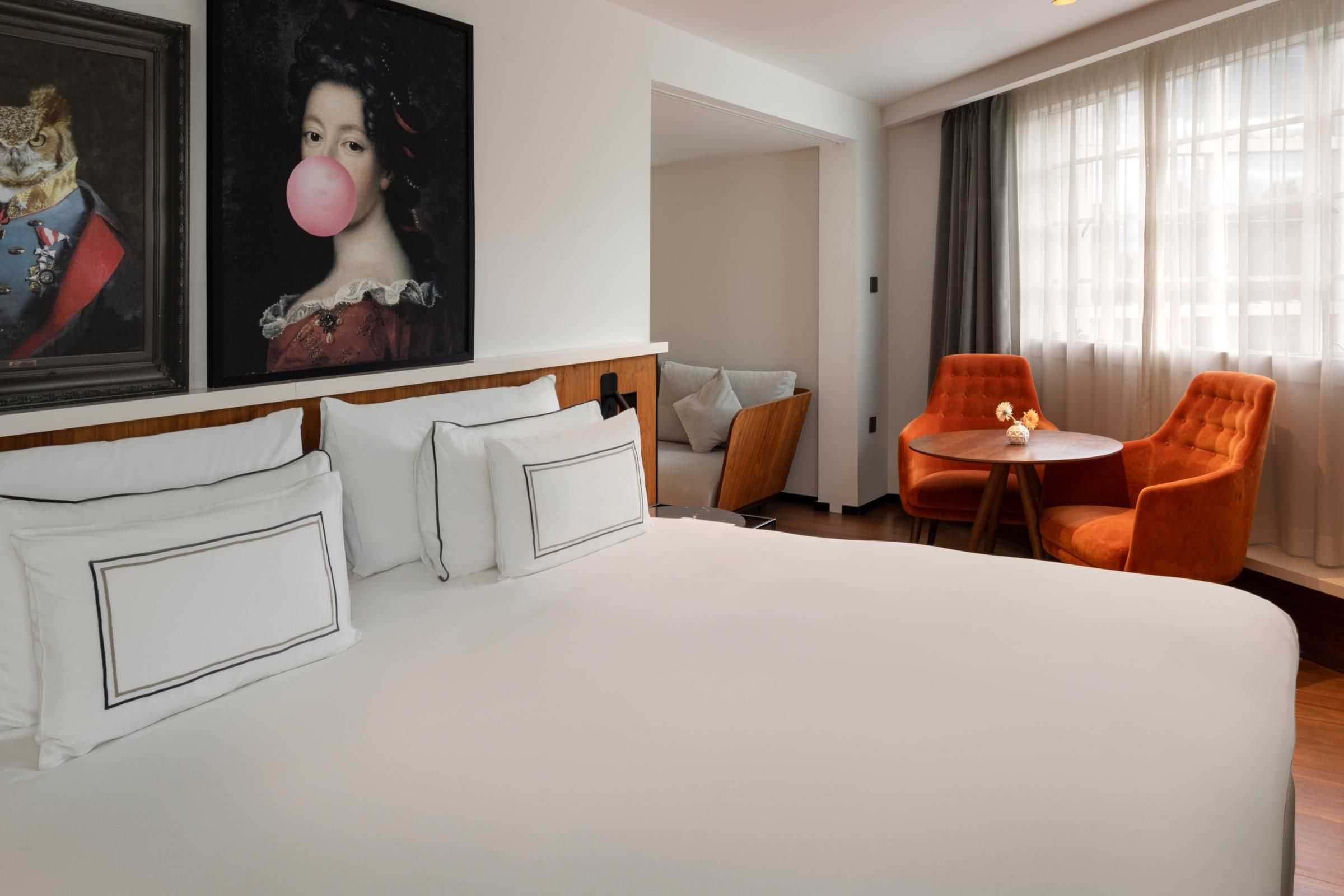 A hotel room with a large bed, two orange chairs, a small table, and a painting on the wall.