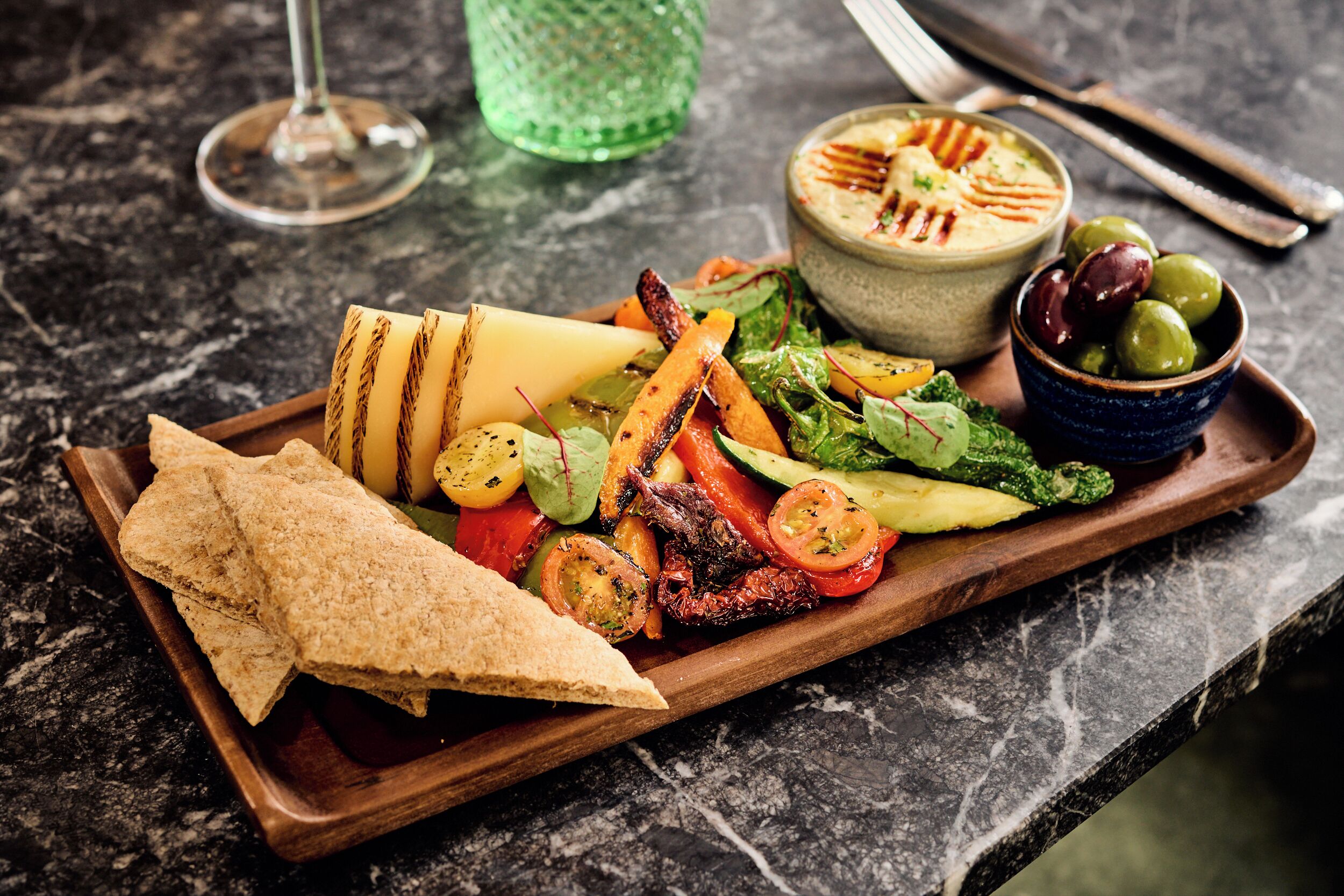 A wooden serving tray with grilled vegetables, a bowl of hummus, olives, and pita bread on a marble countertop.