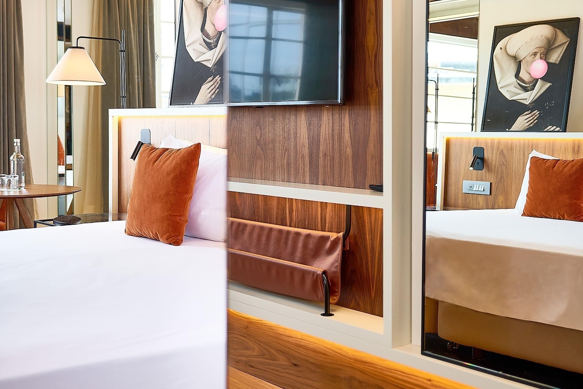 A hotel room with a bed, bedside table, lamp, and a large mirror reflecting the room.