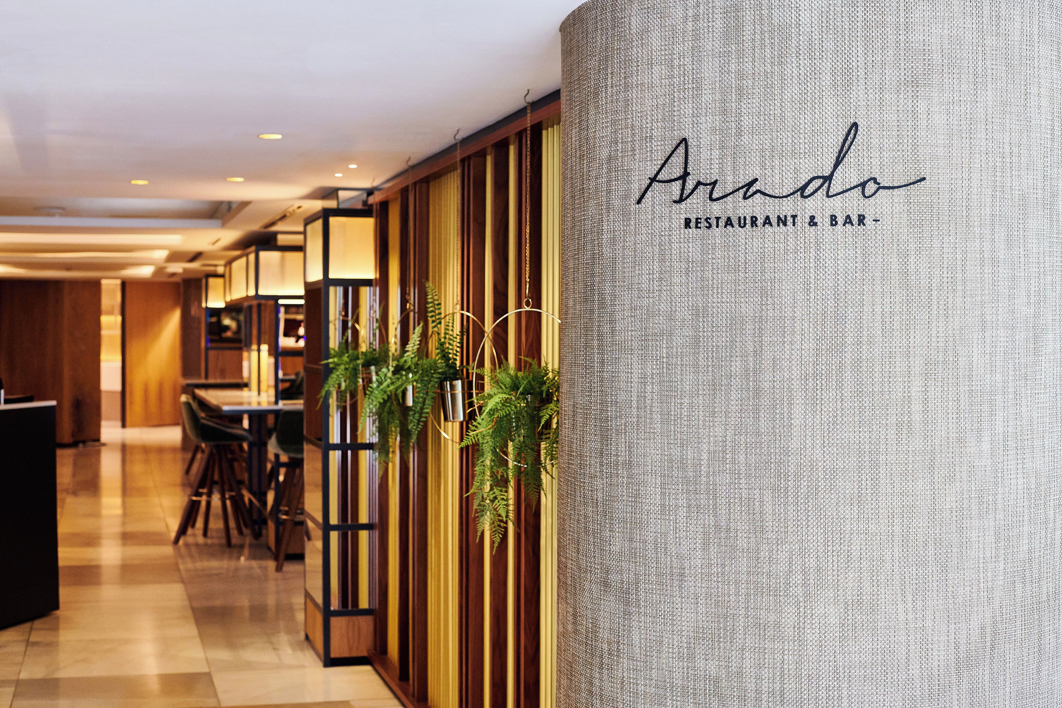 Interior de un restaurante con un panel de madera, plantas colgantes y un letrero que dice 'Arado Restaurant & Bar'.
