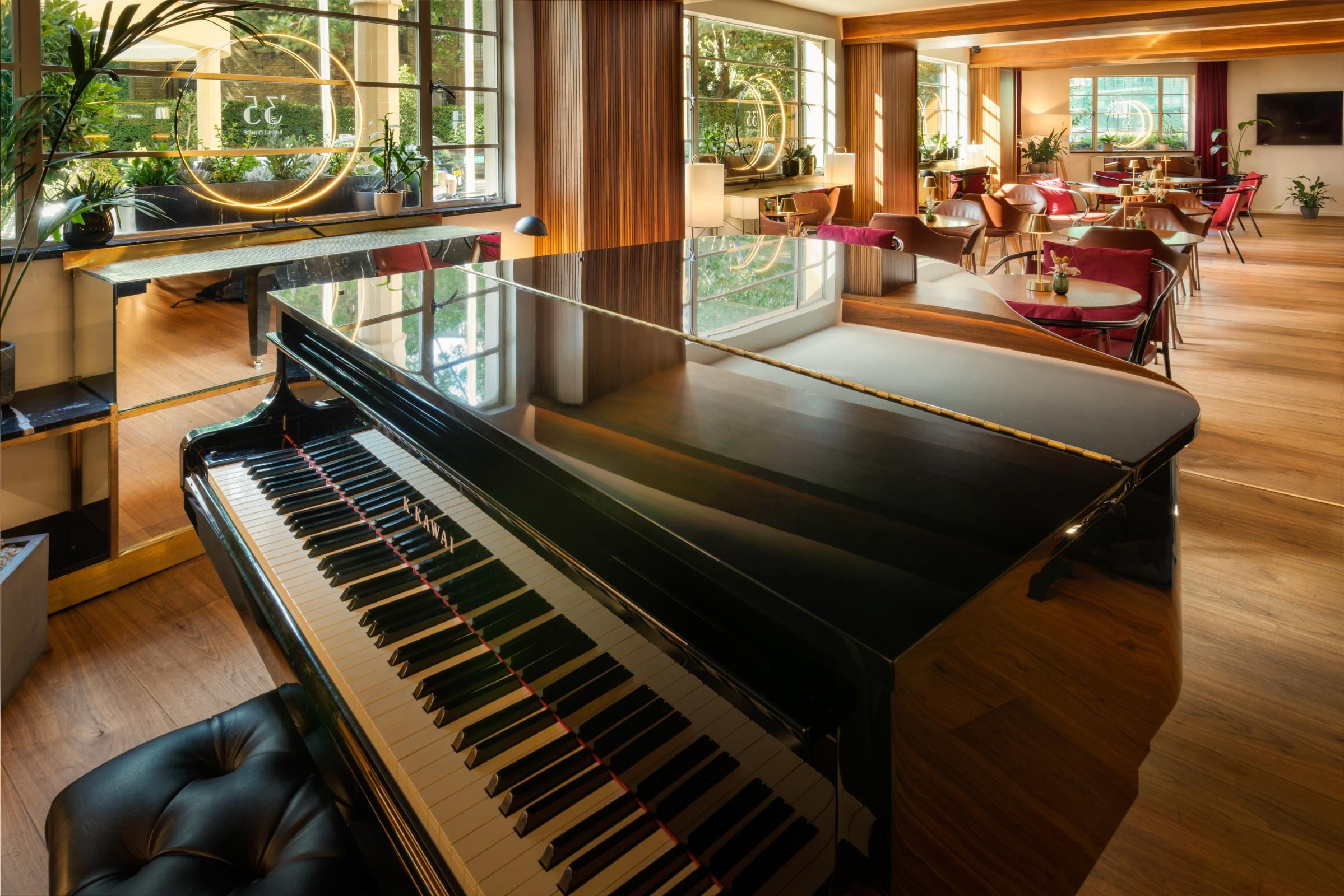 A grand piano in a well-lit room with large windows, a seating area with tables and chairs, and a view of greenery outside.