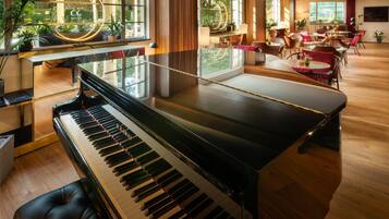 A grand piano in a well-lit room with large windows, a seating area with tables and chairs, and a view of greenery outside.