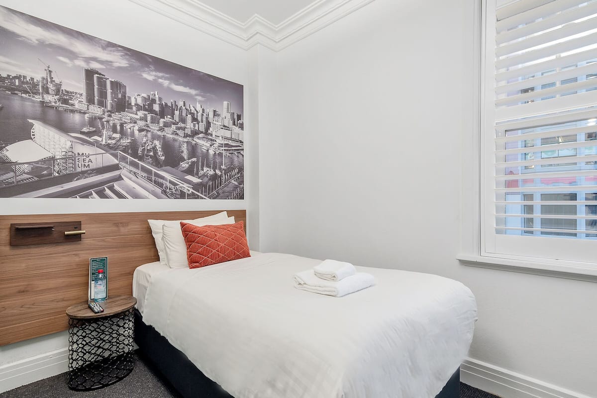 Habitación de hotel con cama, mesita de noche, control remoto, almohada roja, colcha blanca y un póster de un paisaje urbano en la pared.