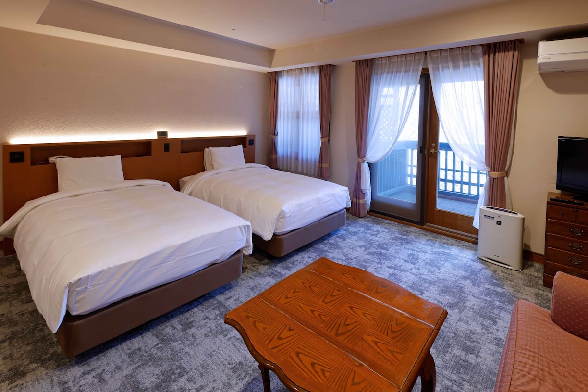 A hotel room with two beds, a wooden table, a television, and a balcony with curtains.