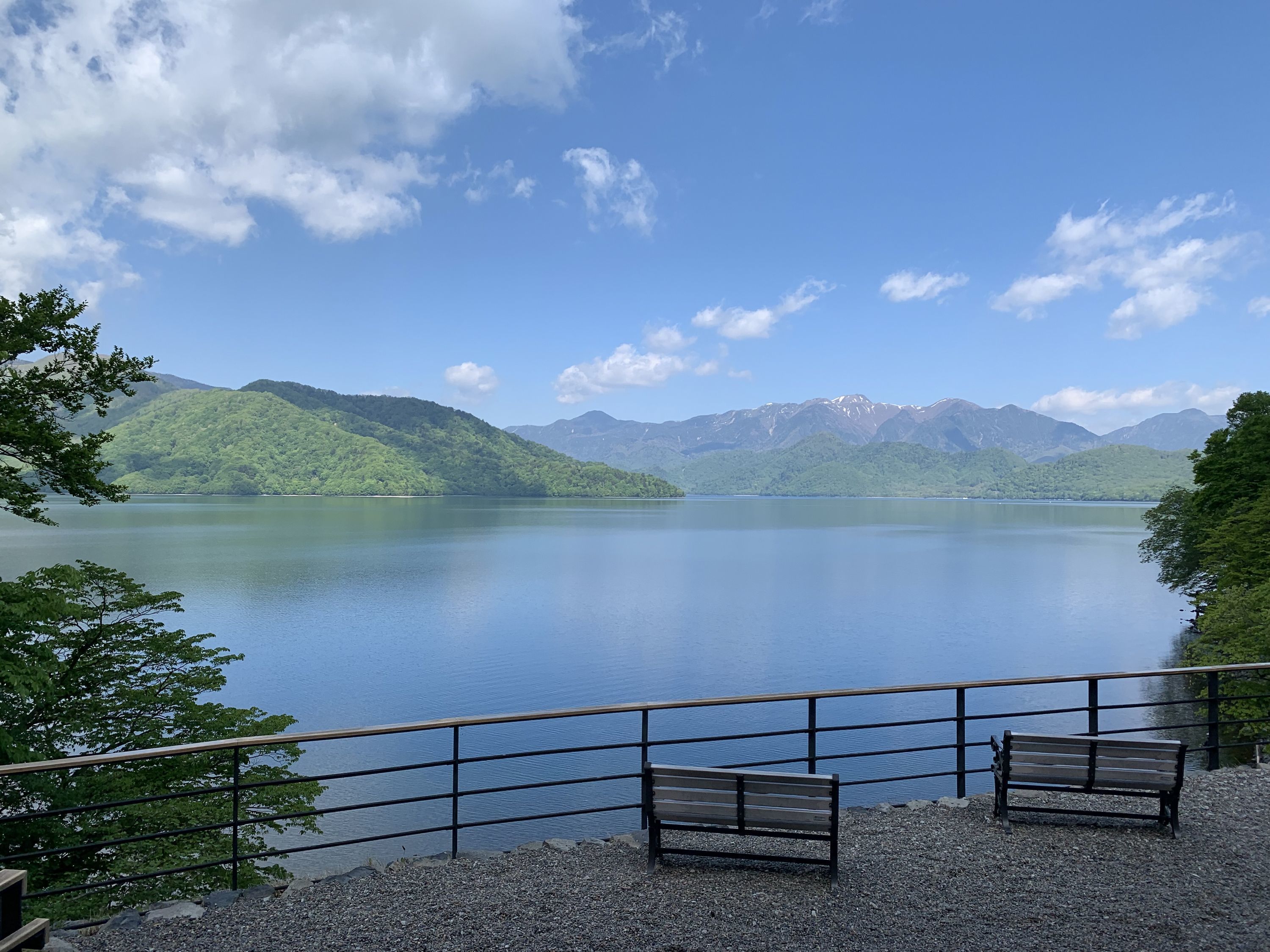 A lakeside view with a bench and railing, surrounded by mountains and trees.