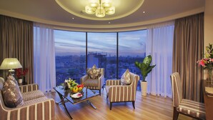 Ein geräumiges Wohnzimmer mit großem Fenster und Blick auf die Stadt, einer runden Deckenleuchte sowie einem Esstisch, der mit Früchten und einer Blumenvase gedeckt ist.