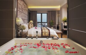 Une chambre d’hôtel avec un grand lit, une baignoire remplie de pétales de roses, une vue sur la ville et un coin repas avec une table et des chaises.
