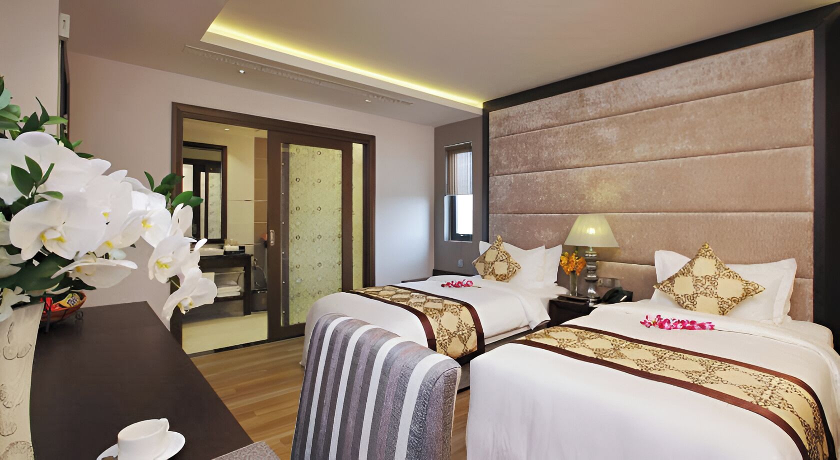 A hotel room with two beds, a nightstand, a lamp, a vase with flowers, and a view of a bathroom.