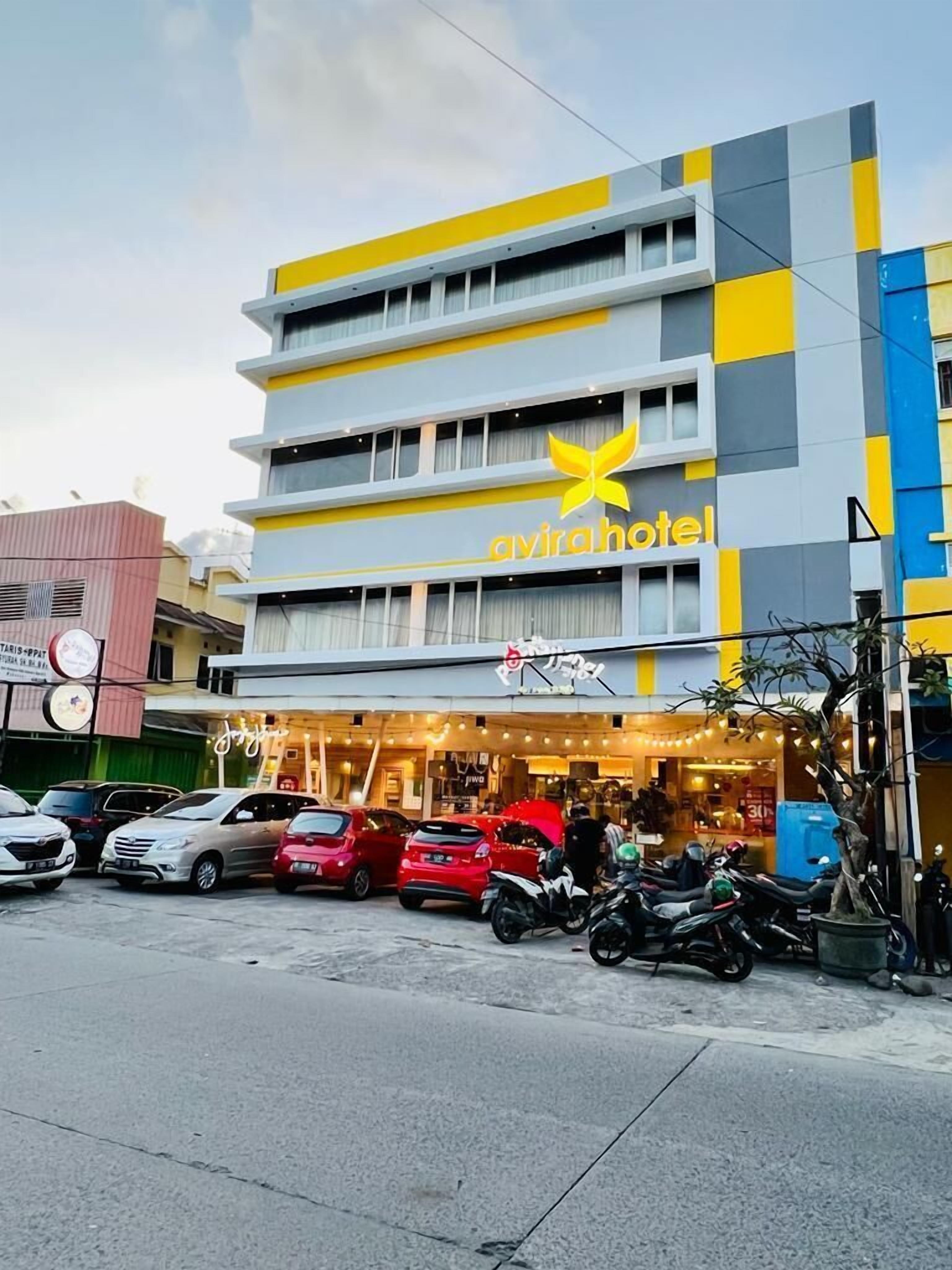 Un hôtel à plusieurs étages, doté d’une façade jaune et grise, d’une enseigne bien visible indiquant « Avira Hotel », et d’un restaurant situé au rez-de-chaussée avec une terrasse.