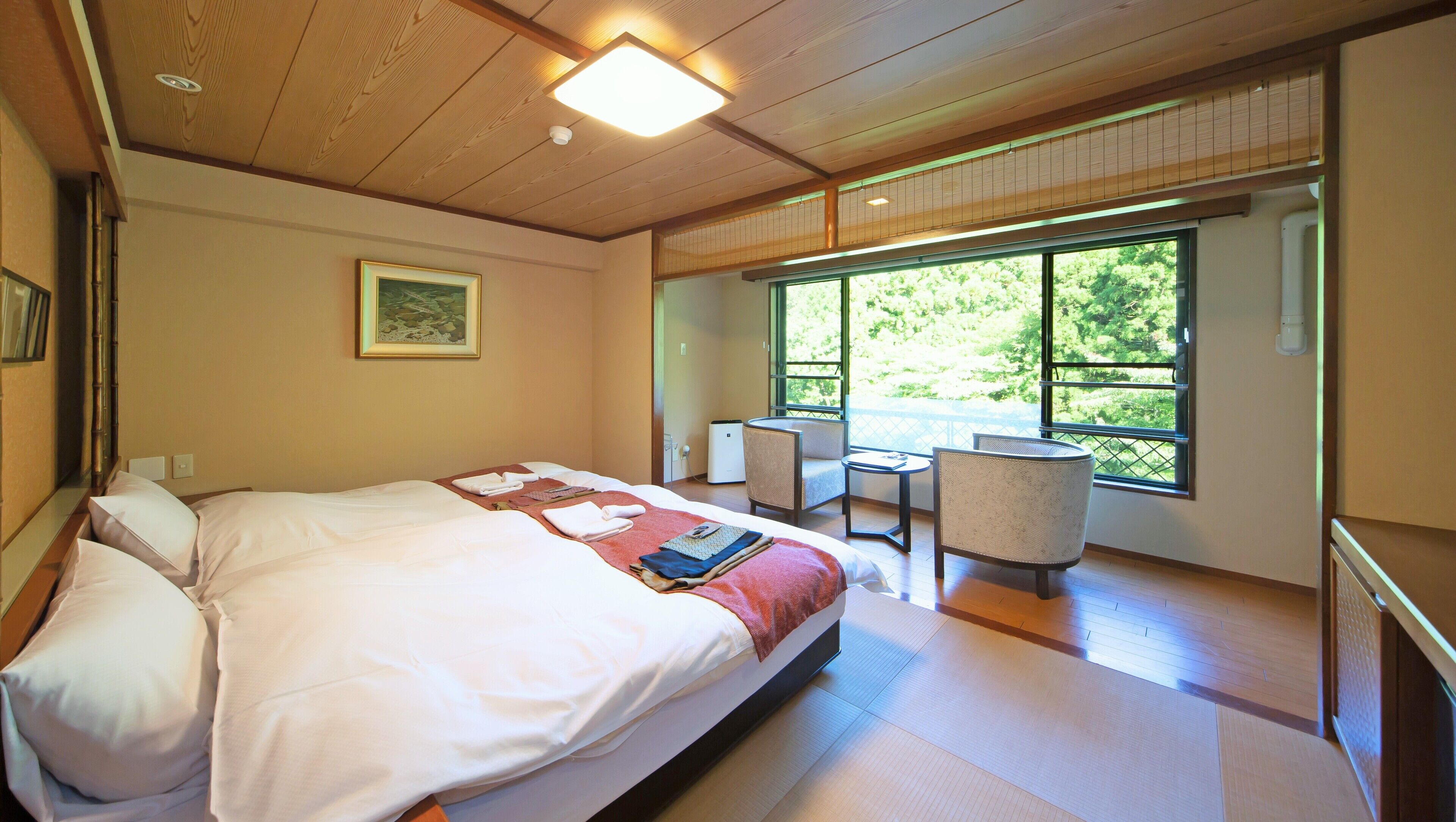A hotel room with a bed, a chair, a table, and a large window with a view of greenery.