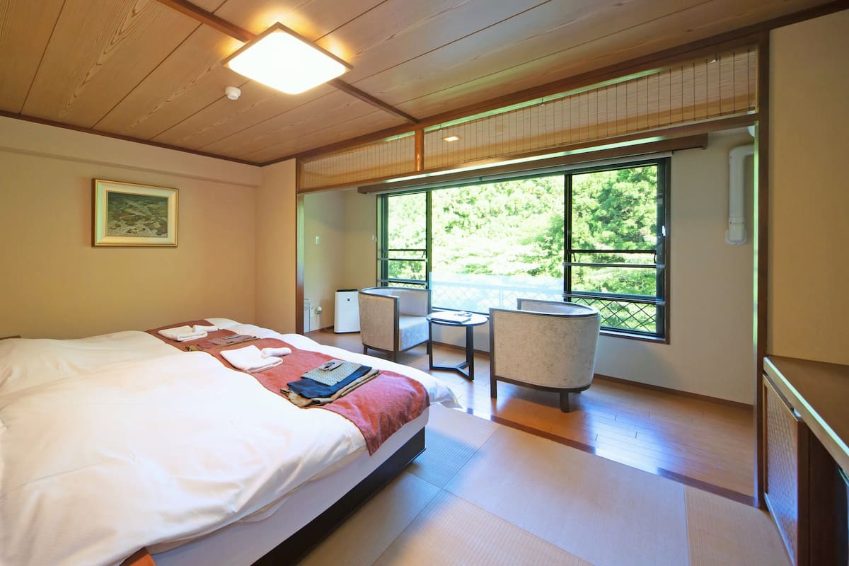 A hotel room with a bed, a chair, a table, and a large window with a view of greenery.