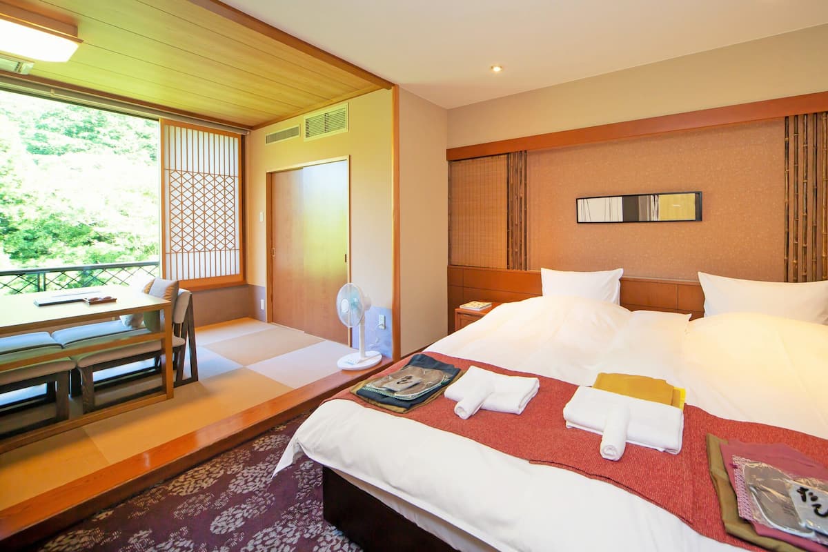 A hotel room with a bed, bedside table, chair, and a view of greenery outside.