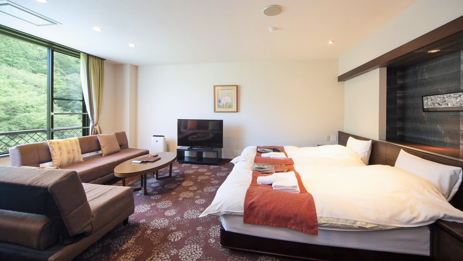A hotel room with a bed, sofa, TV, and a view of greenery.