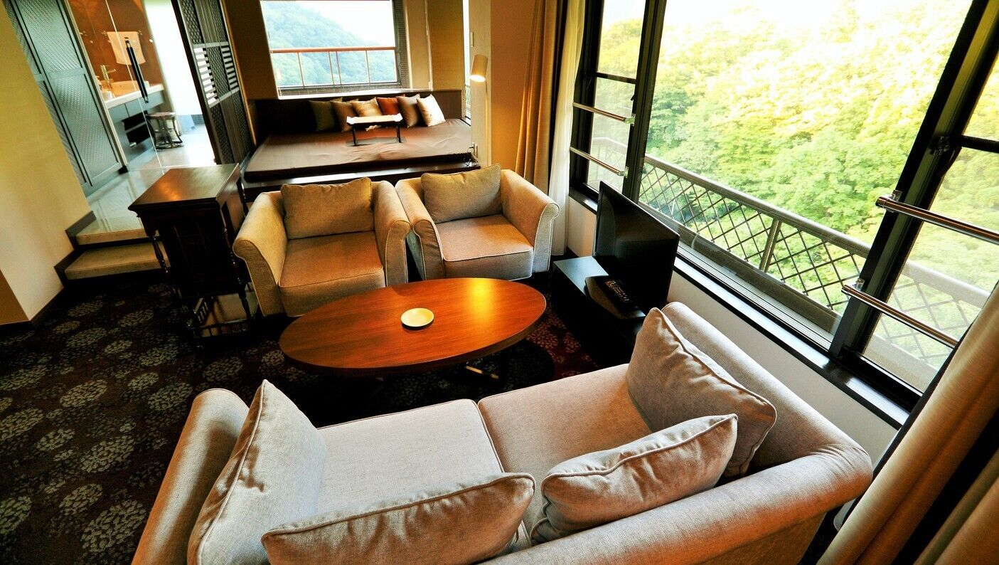 A modern living room with a sofa set, a coffee table, and a view of a green landscape through large windows.