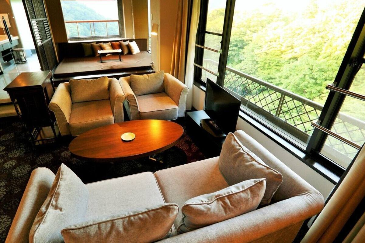Un salon moderne avec un ensemble de canapés, une table basse et une vue sur un paysage verdoyant grâce à de grandes fenêtres.