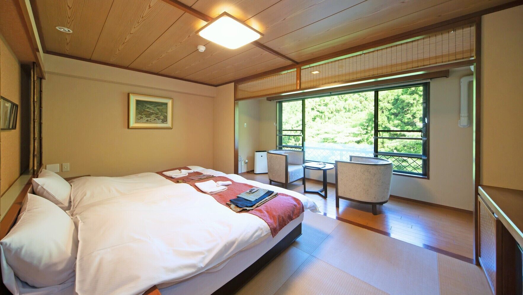 A hotel room with a bed, a small table, two chairs, a window with a view of greenery, and a ceiling light fixture.