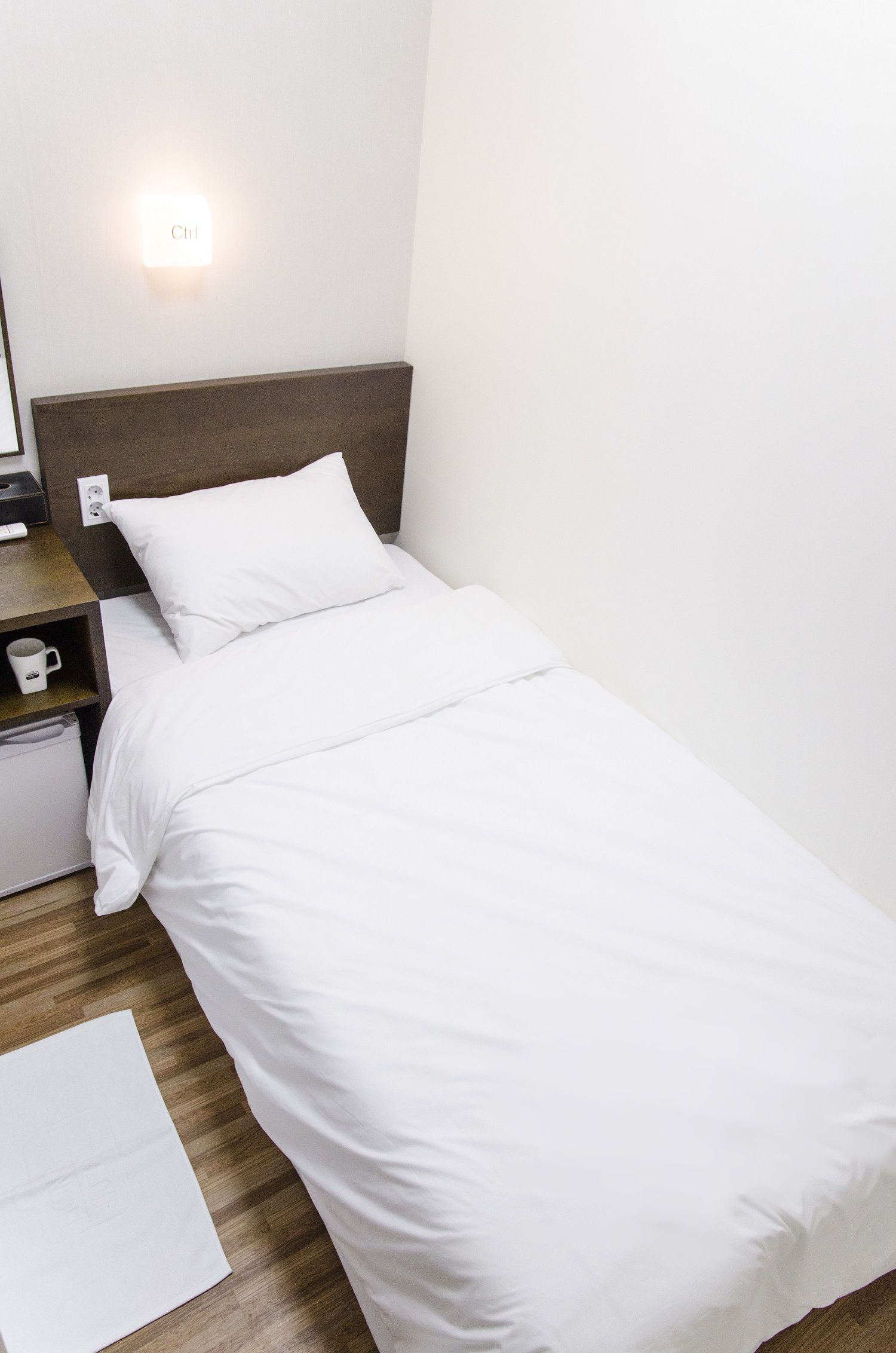 A neatly made bed with white bedding, a bedside table with a cup and a lamp, and a wall-mounted light.
