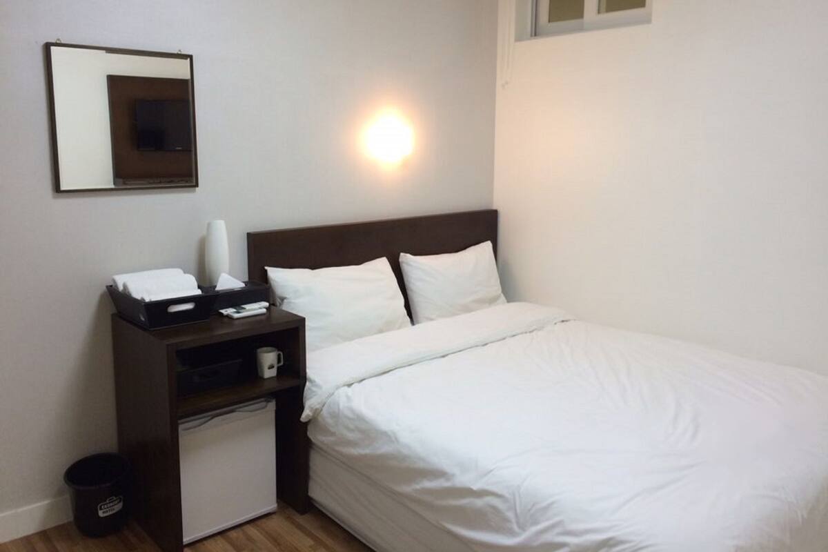 Habitación de hotel con cama, mesita de noche con lámpara, escritorio pequeño con computadora y un cesto de basura.