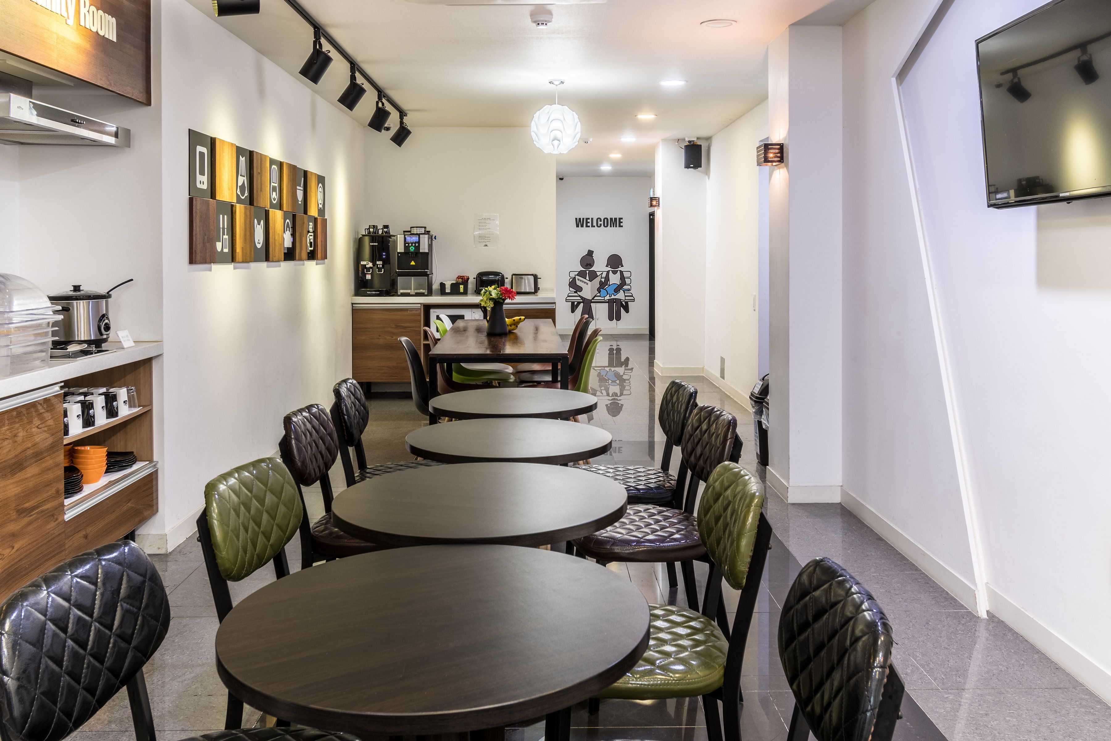 A modern dining area with round tables and cushioned chairs, a kitchen in the background, and a welcome sign on the wall.
