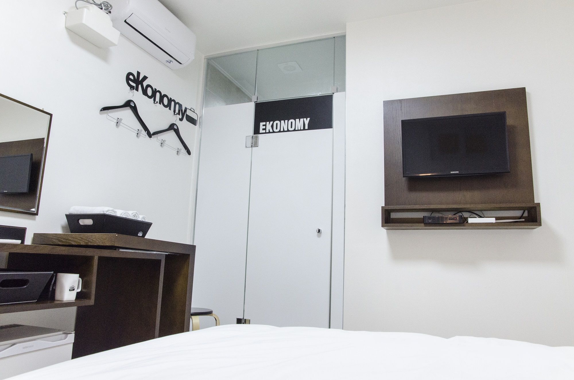 A hotel room with a bed, a desk with a computer, a TV mounted on the wall, and a wardrobe with a sign that reads "EKONOMY".