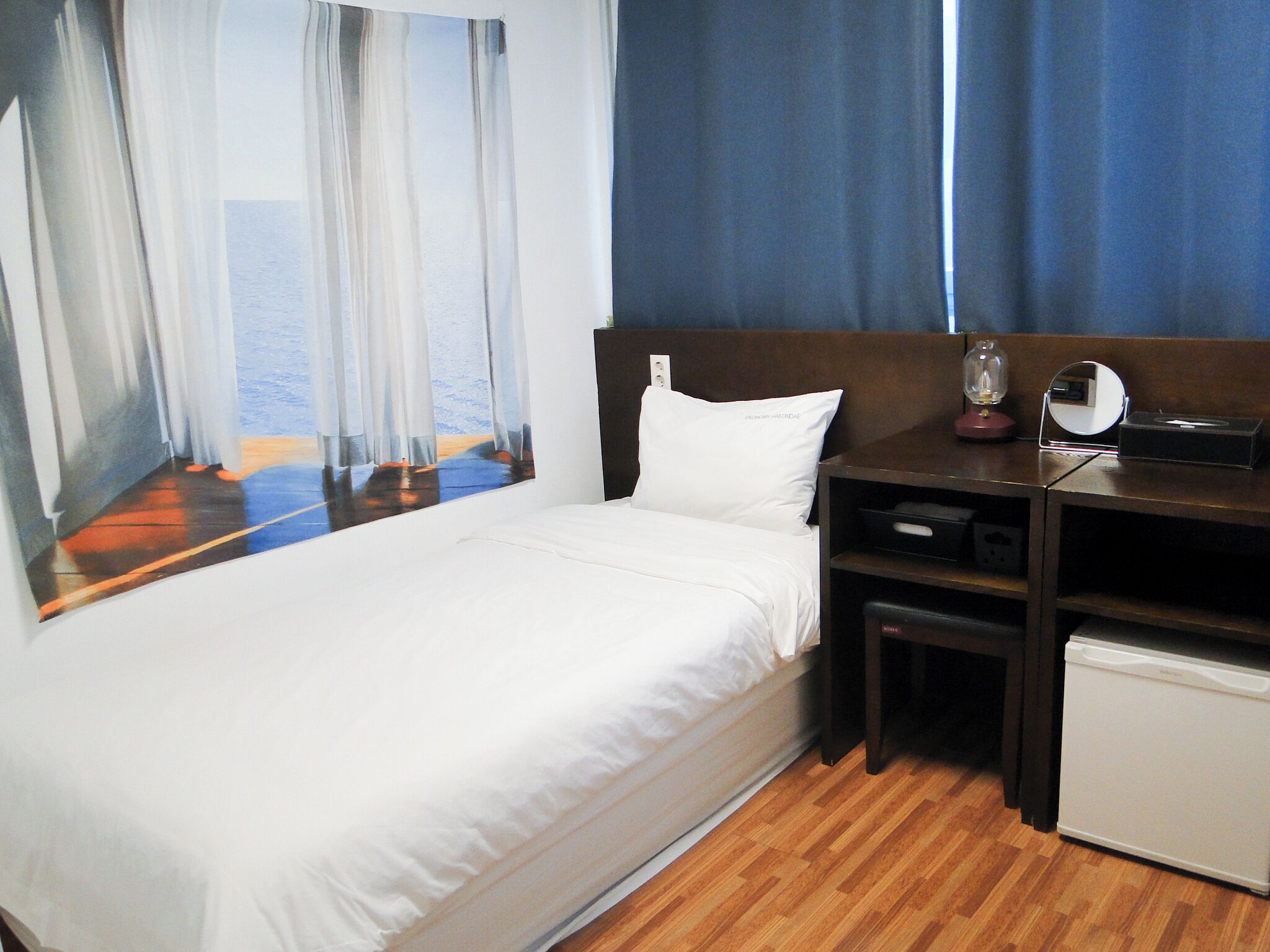 A hotel room with a bed, a desk with a mirror, and a view of the sea through sheer curtains.