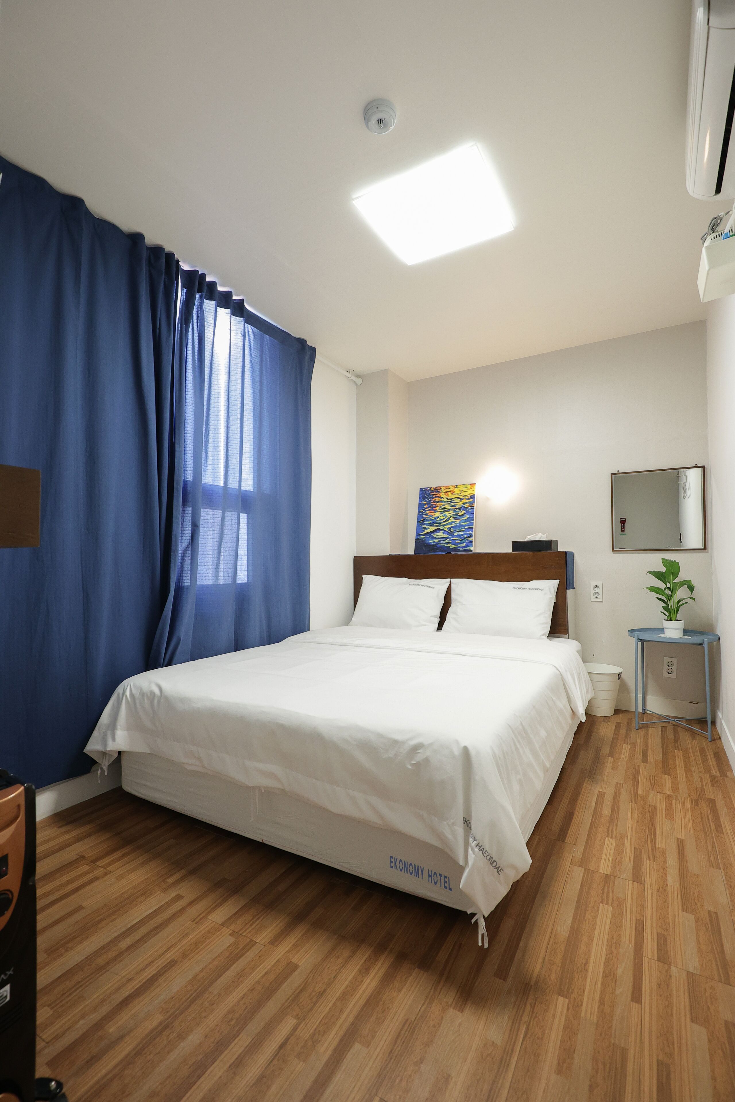 A hotel room with a wooden floor, a bed with white bedding, a nightstand with a plant, a painting on the wall, and a ceiling light.