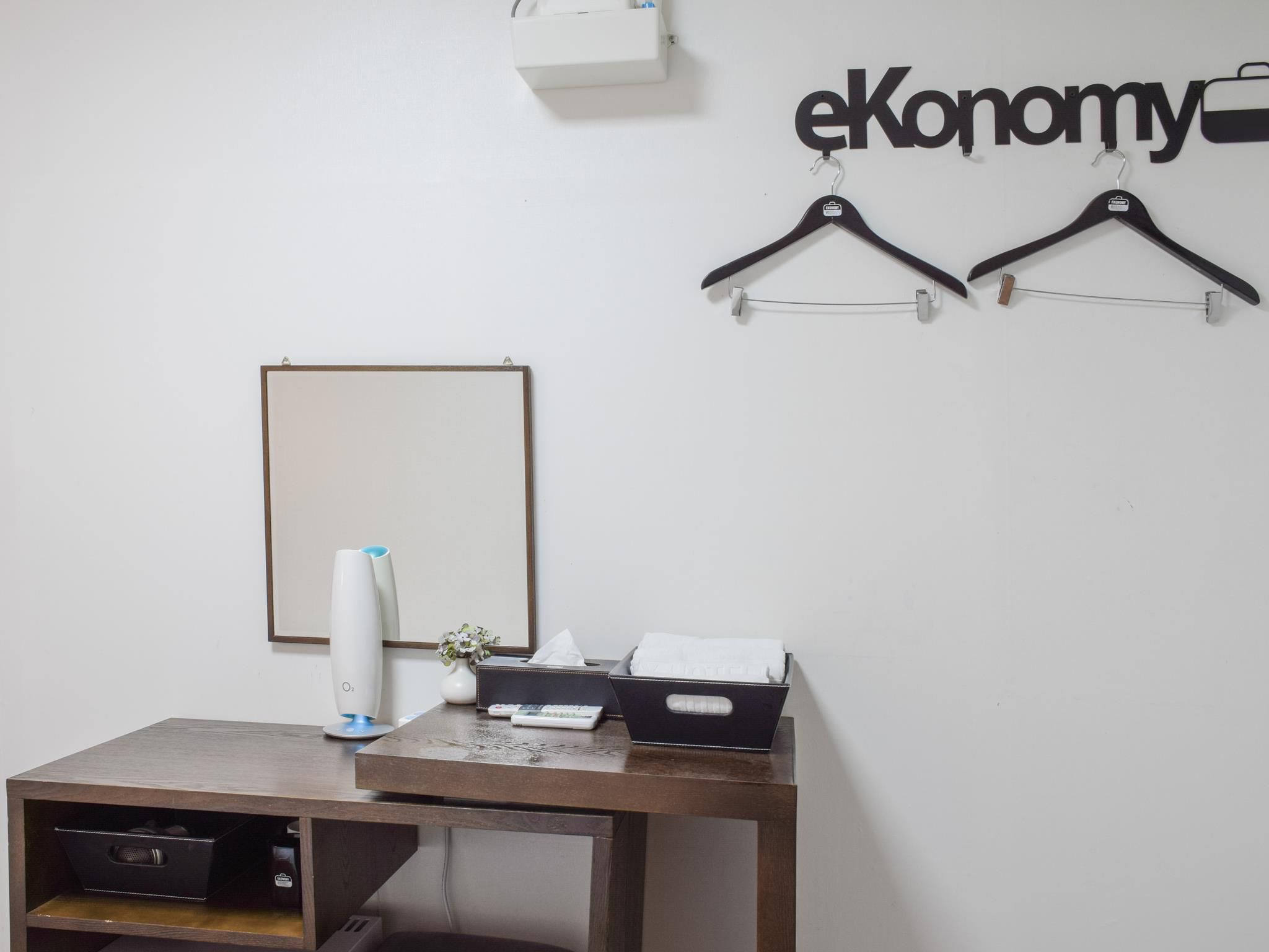 Ein Zimmer mit Schreibtisch aus Holz, einer weißen Vase, einer Taschentuchbox und zwei schwarzen Kleiderbügeln an der Wand.