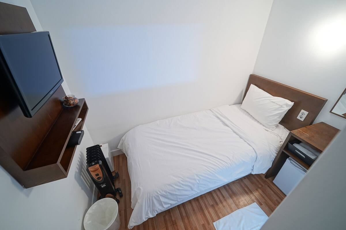 Una habitación de hotel pequeña y ordenada, con una cama, un televisor, una mesita y un cubo de basura.