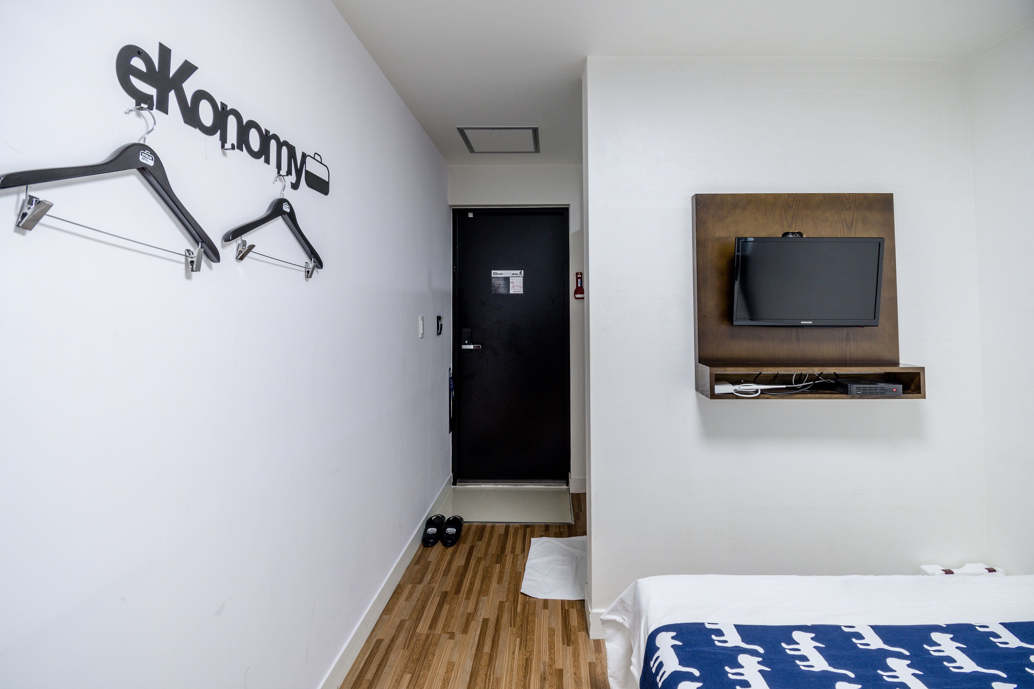A hotel room with a bed, a TV mounted on the wall, a coat rack, and a door leading to the hallway.