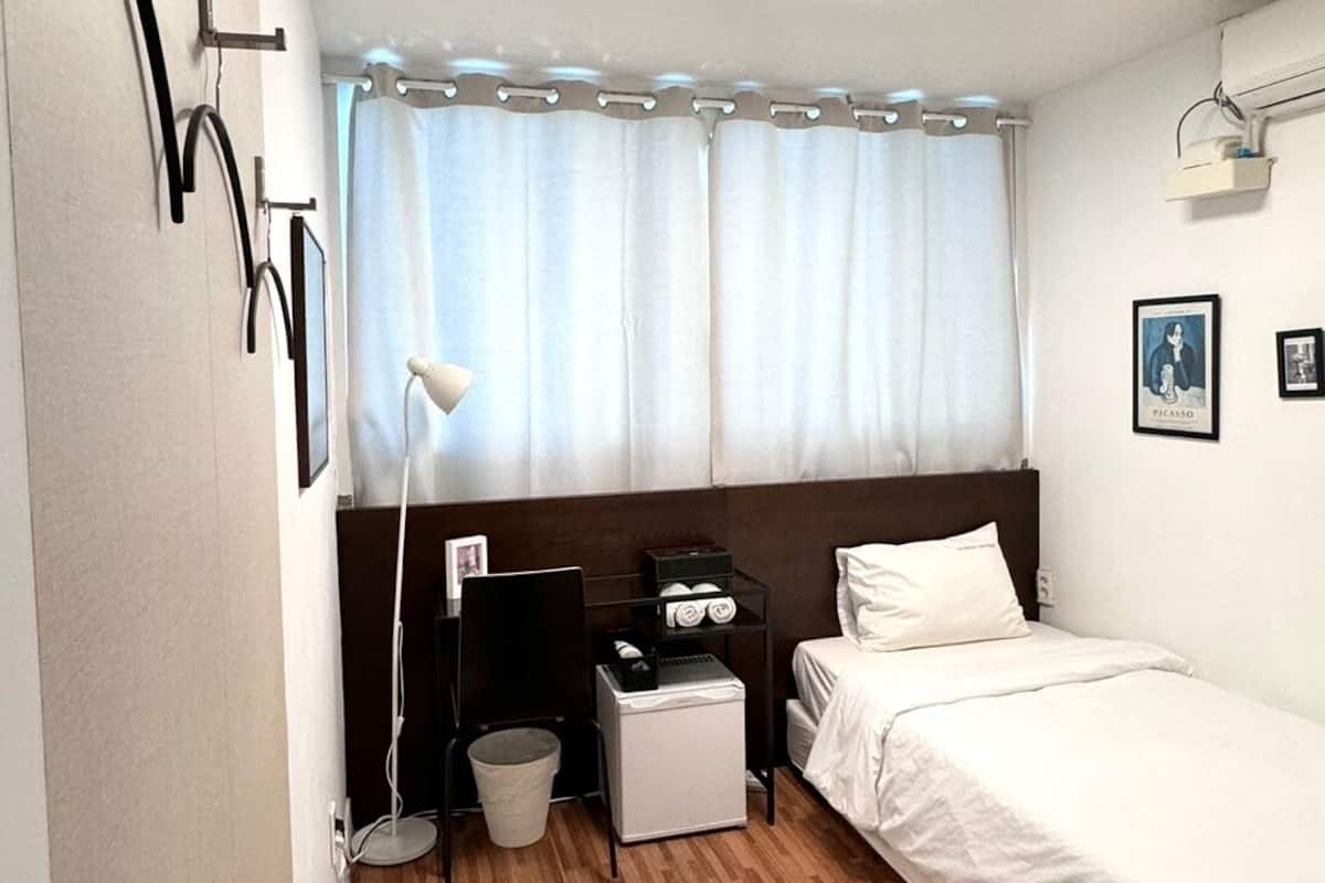Habitación de hotel con piso de madera, una cama, una mesita de noche, un escritorio con lámpara y una ventana con cortinas.