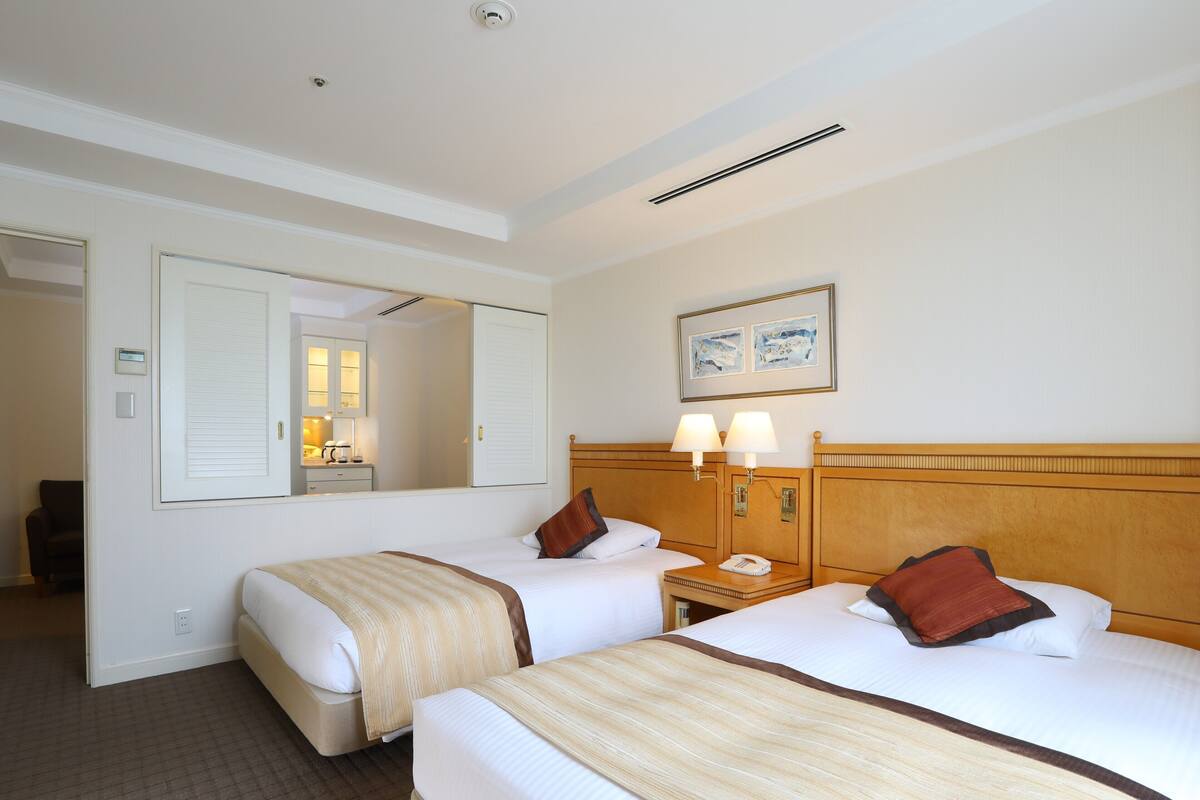 A hotel room with two beds, a bedside table with a lamp, a mirror, and a framed picture on the wall.