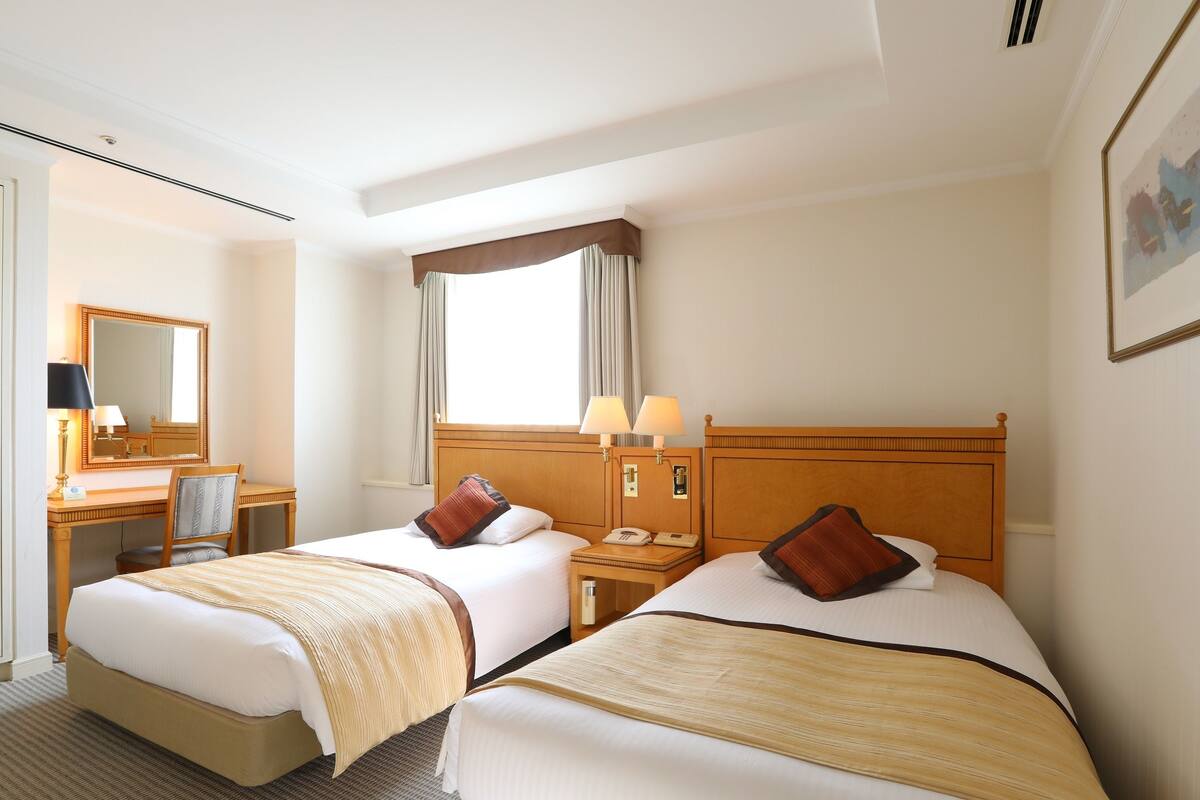 A hotel room with two beds, a desk, a chair, a mirror, and a window with curtains.