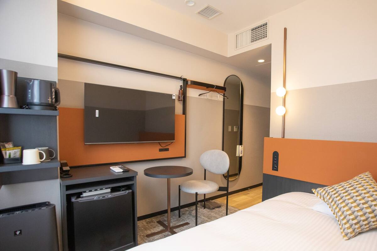 A hotel room with a bed, a desk with a chair, a small table, a mirror, and a TV.