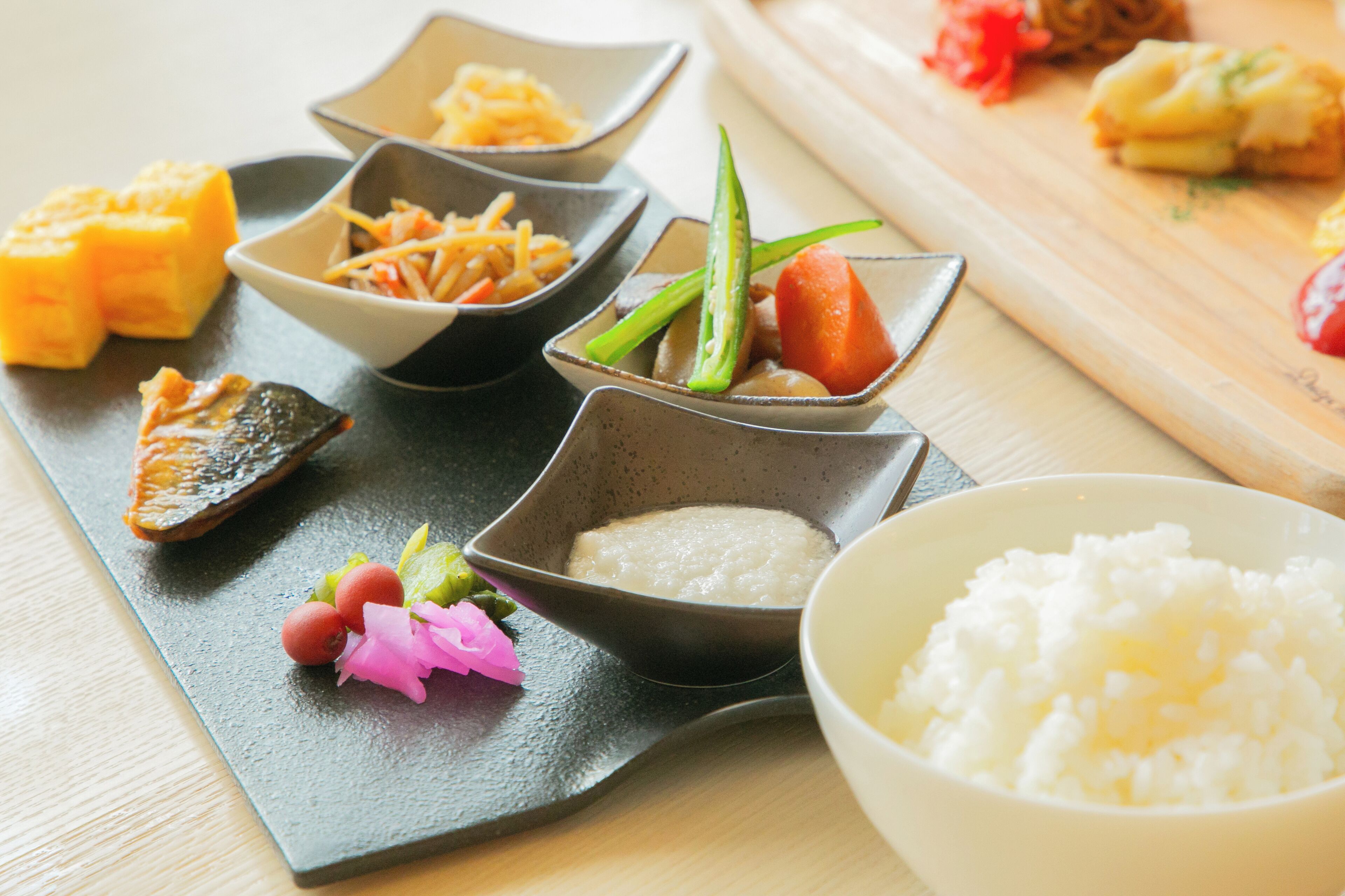 A variety of dishes on a black slate plate, including rice, pickled vegetables, and a piece of fish.