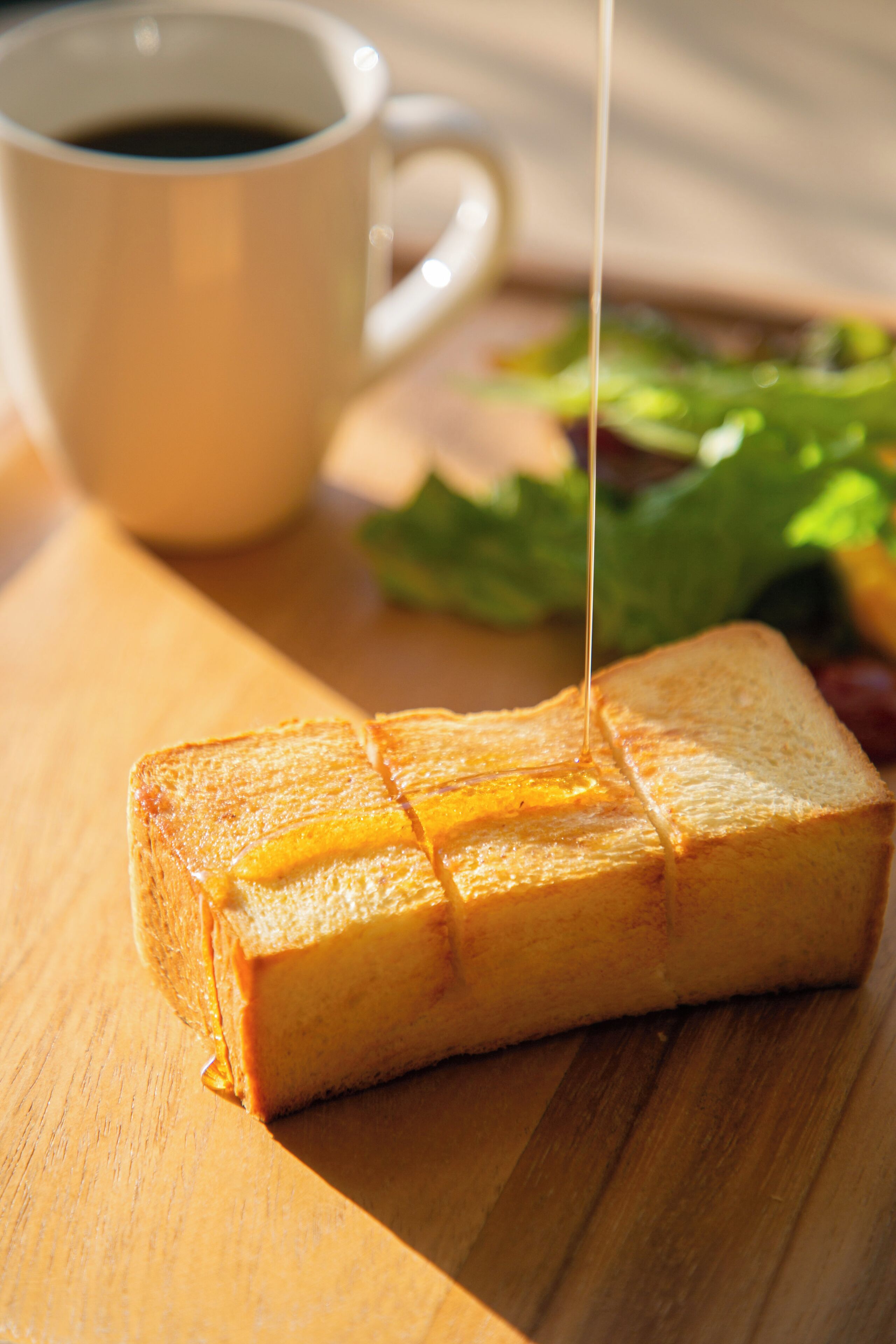 Un sándwich con queso fundido al que se le añade salsa, una taza de café y una ensalada como acompañamiento sobre una mesa de madera.
