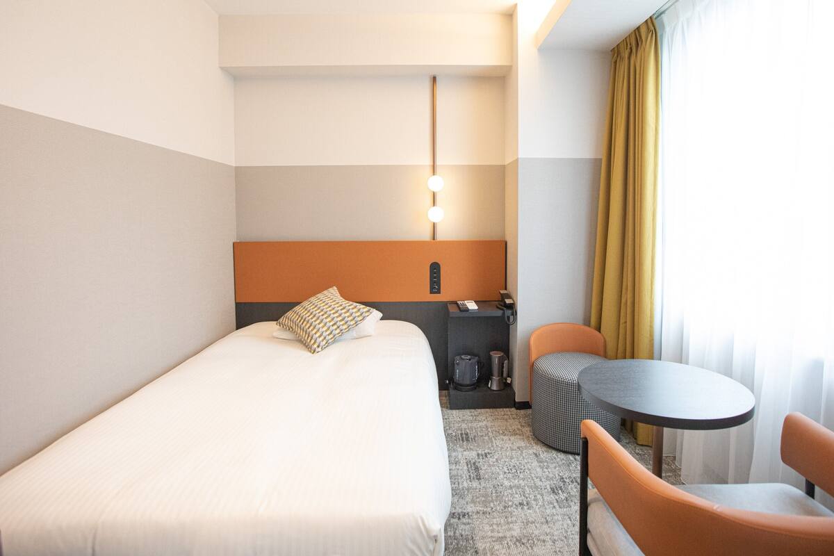 A hotel room with a bed, a desk, a chair, a small table, and a lamp.