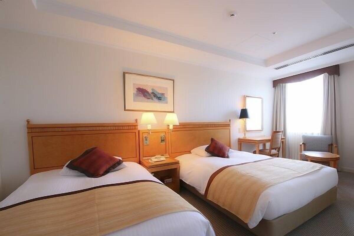 A hotel room with two beds, bedside lamps, a small table, a chair, and a window with curtains.