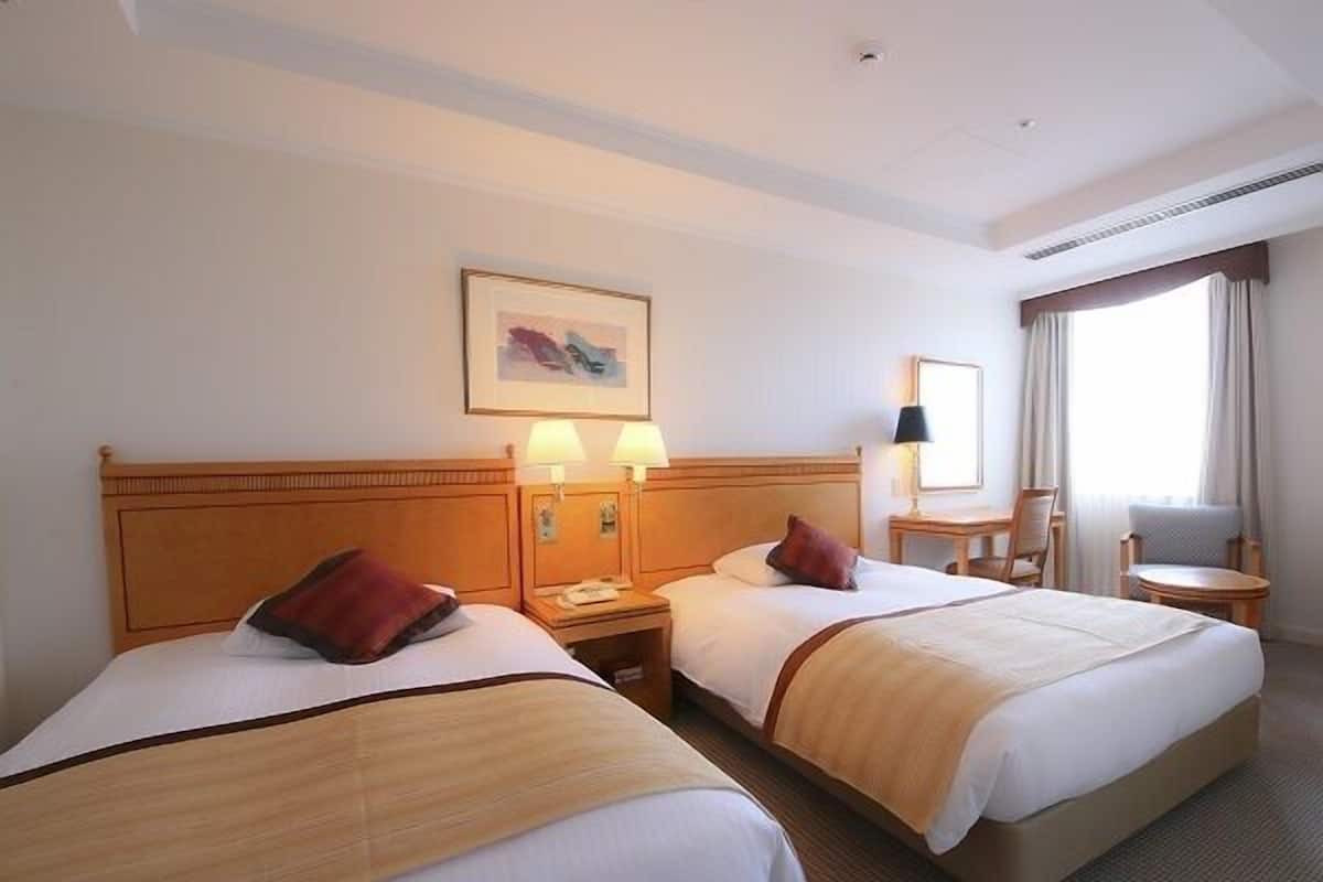 A hotel room with two beds, bedside lamps, a small table, a chair, and a window with curtains.