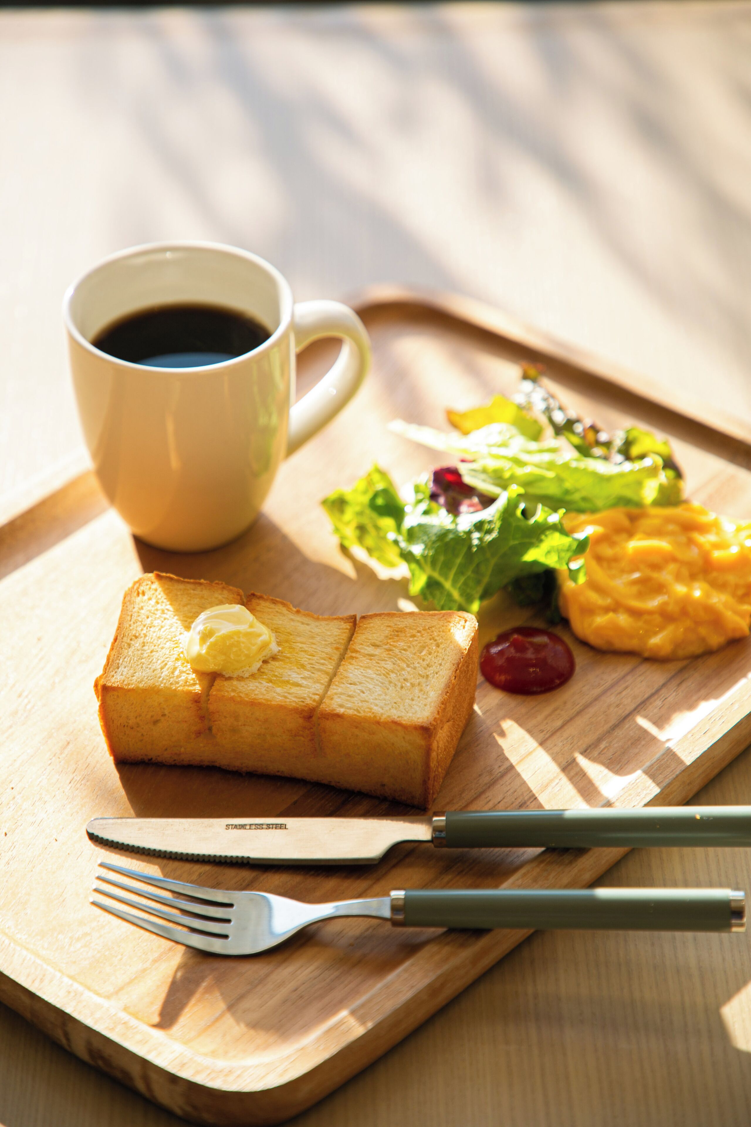 Un plateau de petit-déjeuner avec des toasts, du beurre, une tasse de café, une salade et des œufs brouillés, le tout disposé sur une planche en bois.