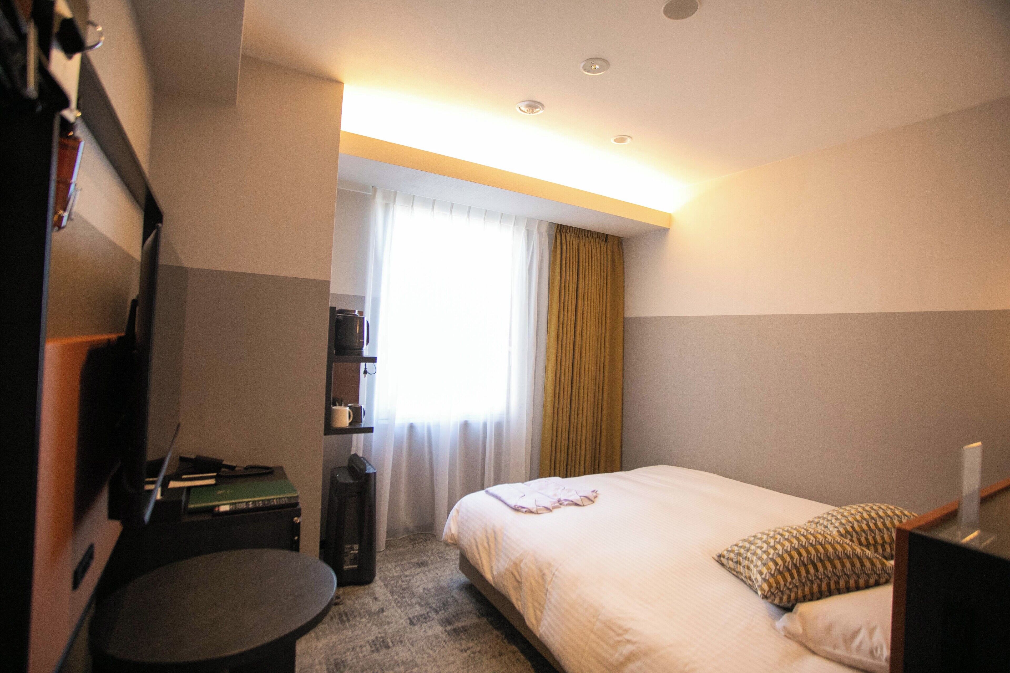 A hotel room with a bed, a desk, a chair, a small table, a wardrobe, and a window with curtains.