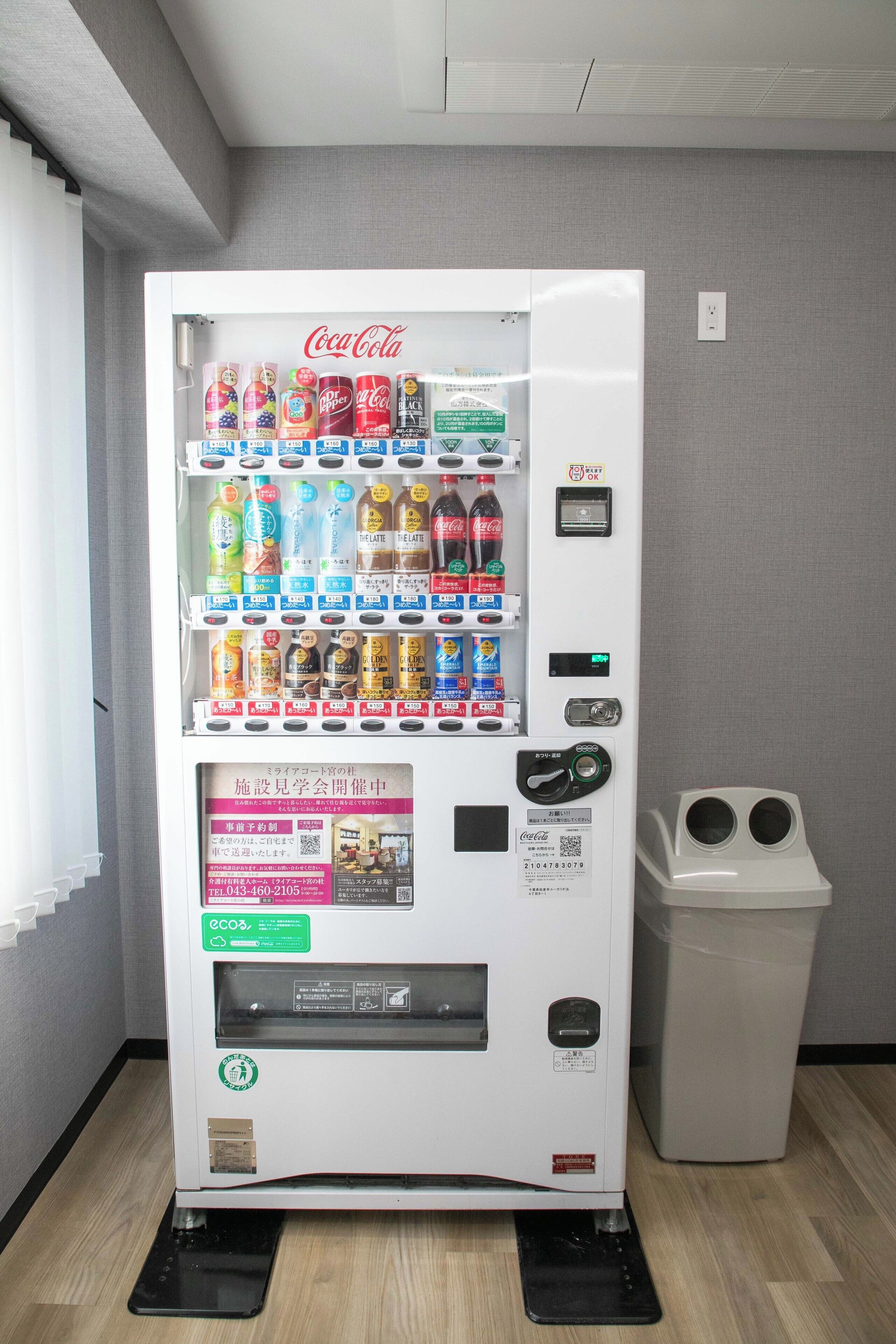 A vending machine with a Coca-Cola logo, various drink options, and a trash bin.