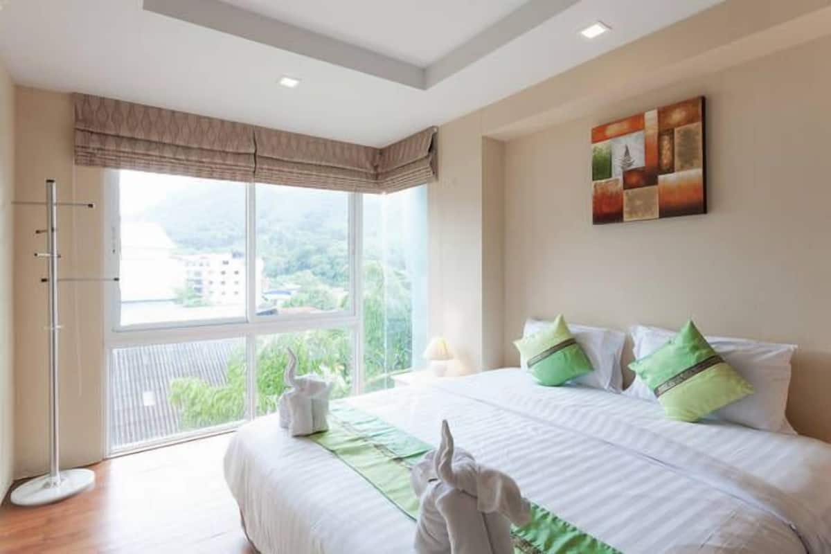 A hotel room with a large window, a bed with green pillows, and a painting on the wall.